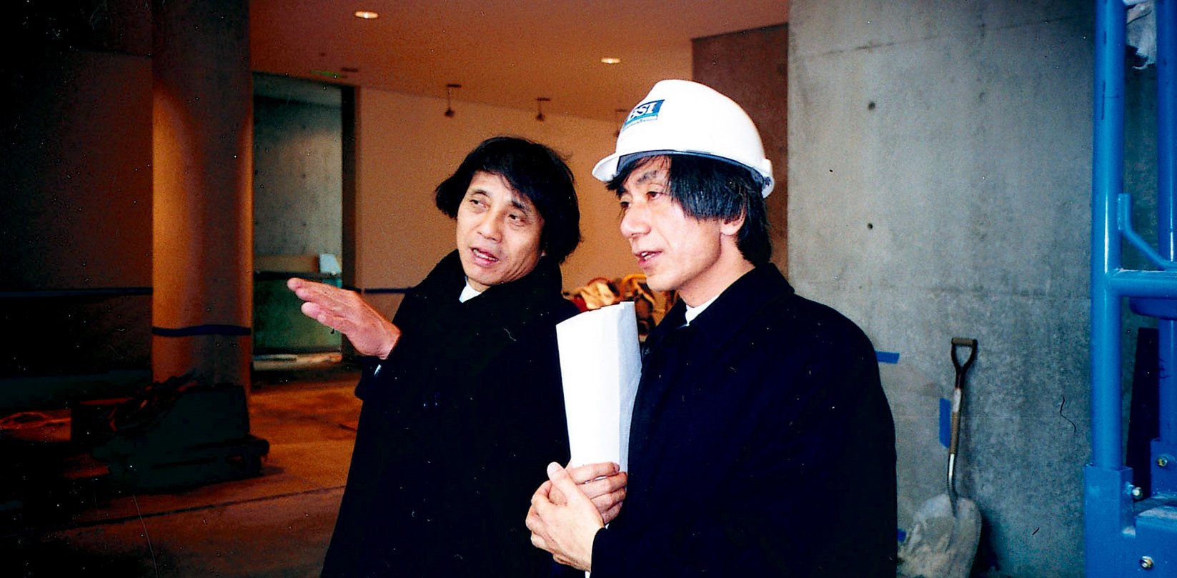 Tadao Ando talks with a construction worker, holding out his hand.