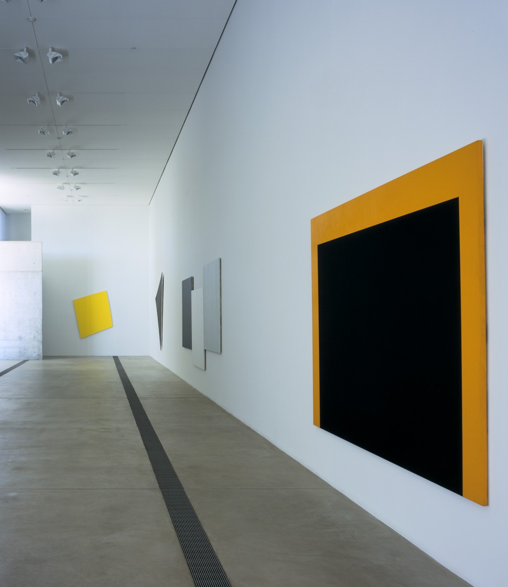 A view of Kelly's work in the Main Gallery's north and east walls, which includes a tilted yellow square, two gray paintings on geometric canvases, and a square black and orange painting.