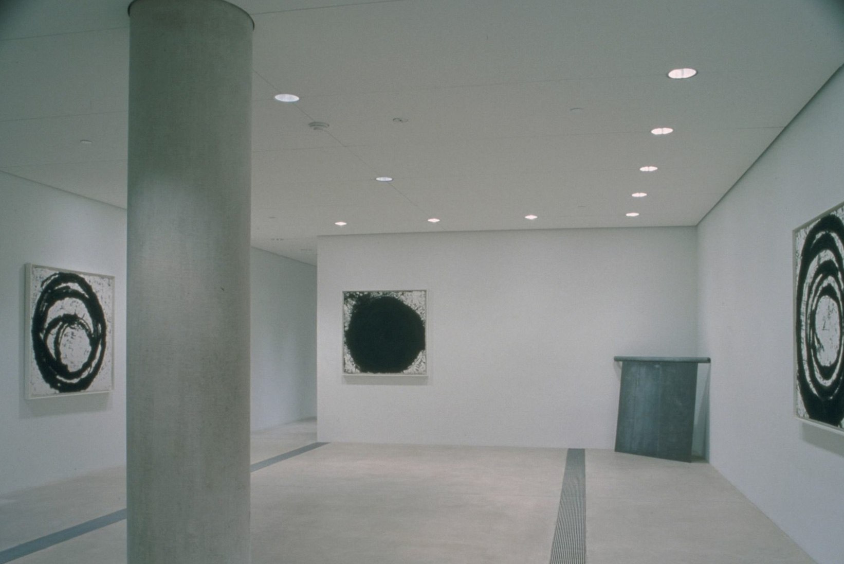 A view of the Entrance Gallery, with three black and white square canvases with circular designs and a steel square that leans against a corner.