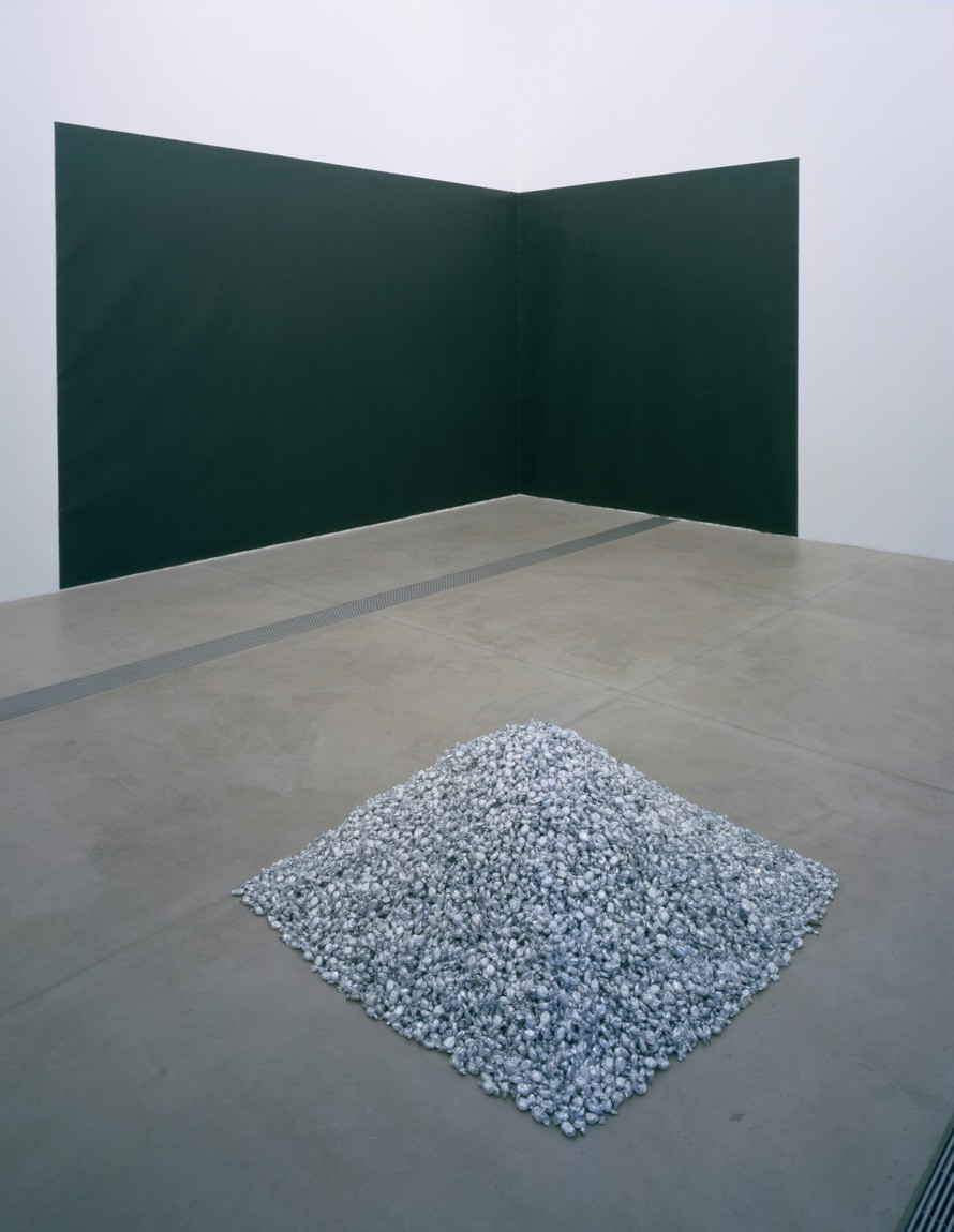 A pile of small gray bits sits on the Cube Gallery floor across from Kelly's "Pacific Judson Murphy," a large black rectangular piece which runs across one wall to another in a corner.