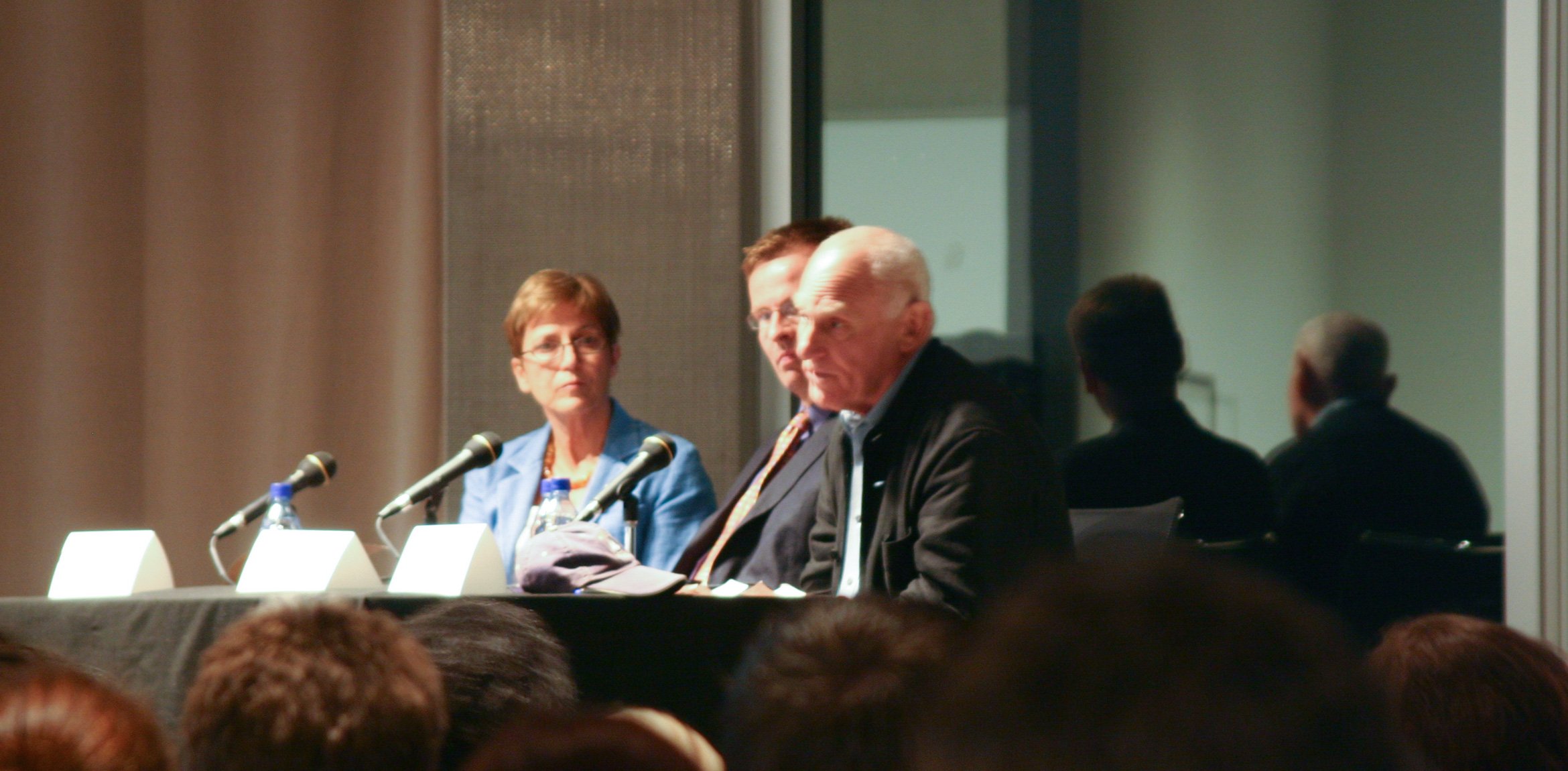 Richard Serra and Carmen Giménez sit at a panel table in front of an audience. Serra speaks into a microphone on the table.