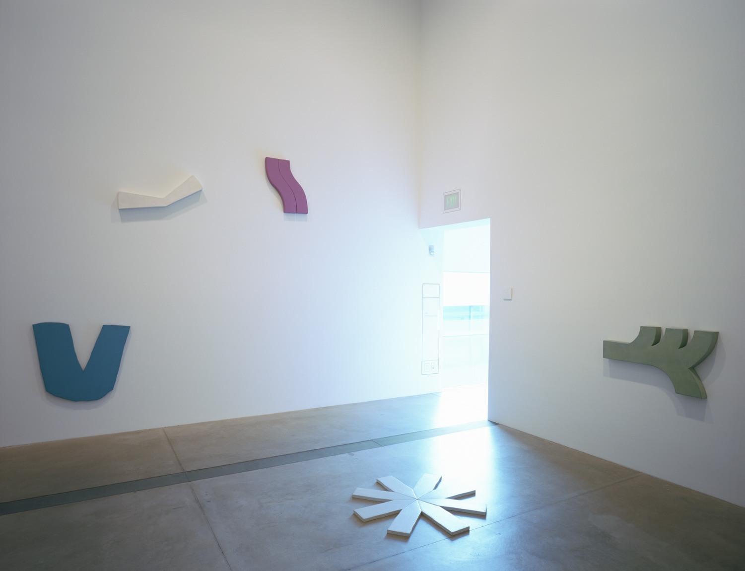 Colorful, flat, wooden sculptures of varying shapes by Richard Tuttle are installed on the walls and floor of the Cube Gallery.