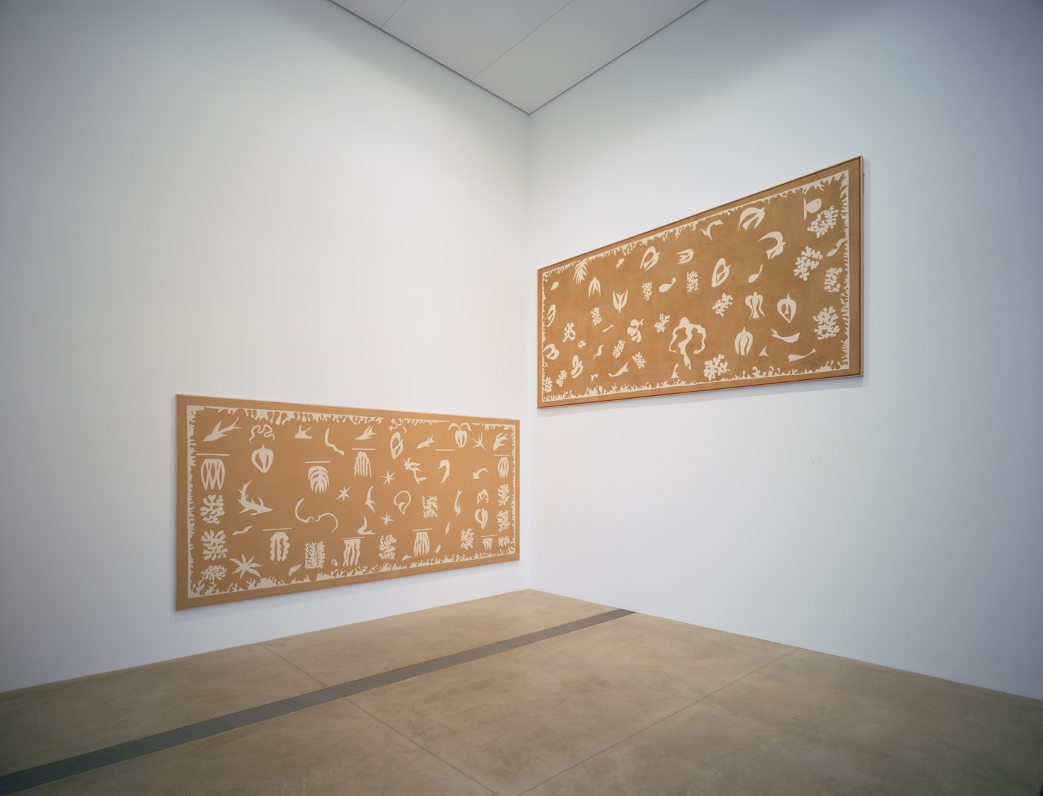 Two Matisse large, wide tan panels decorated with white designs titled "Oceania, the Sea" and "Oceania, the Sky" are each installed on a wall in the Cube Gallery.