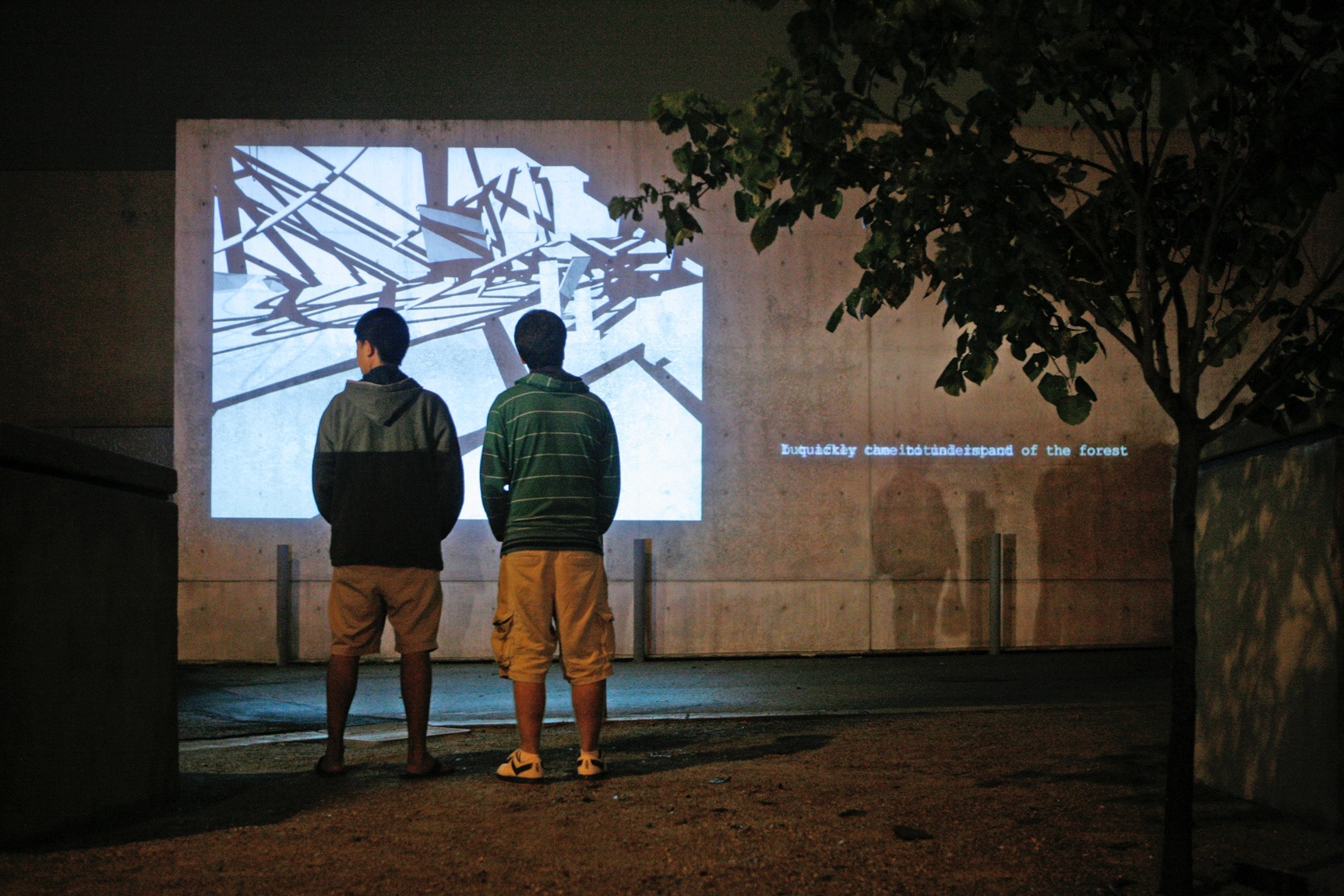 Two visitors stand outside before Ann Lislegaard's film "Crystal World" projected on a concrete wall at night.