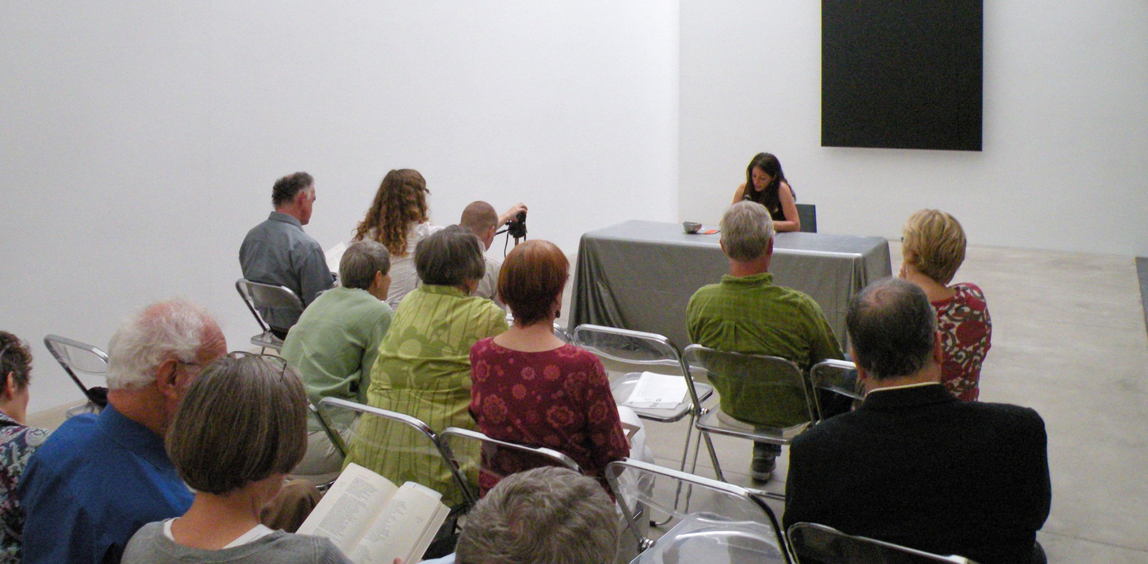 A community member sits at a table in front of Ellsworth Kelly's "Blue Black" and reads from a page into a microphone for an audience.