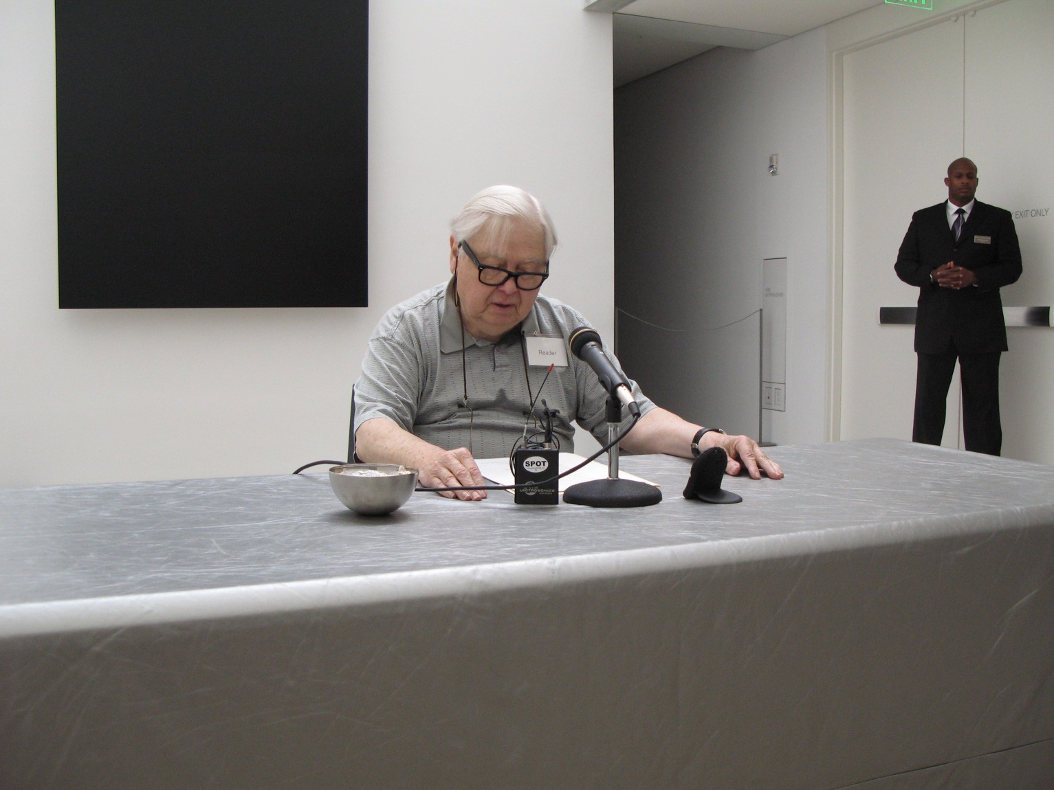 A community member sits at a table in front of Ellsworth Kelly's "Blue Black" and reads from a page into a microphone.