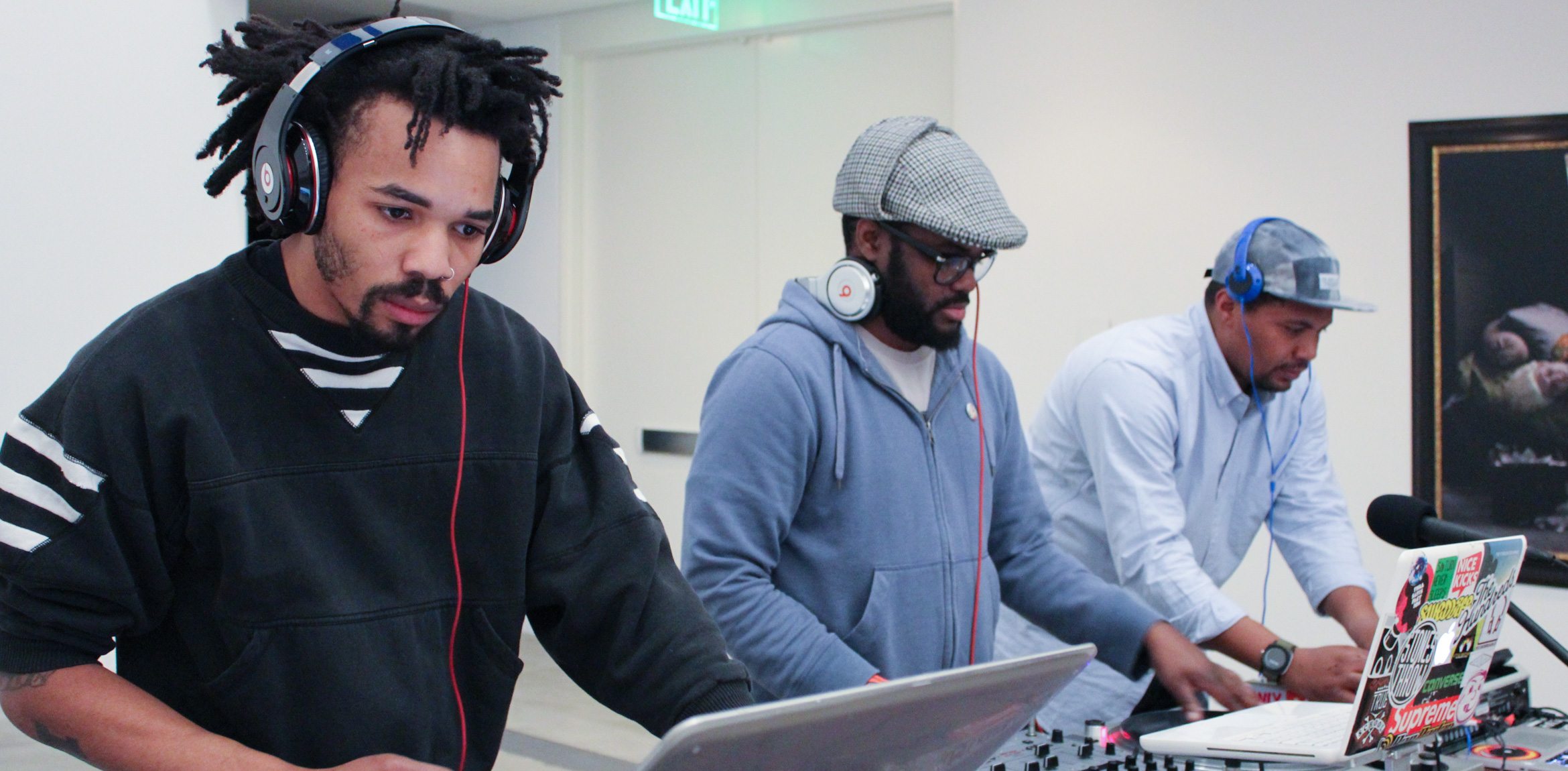 Three DJs wearing headphones are focused on their computers at a table in the Lower-Main Gallery.