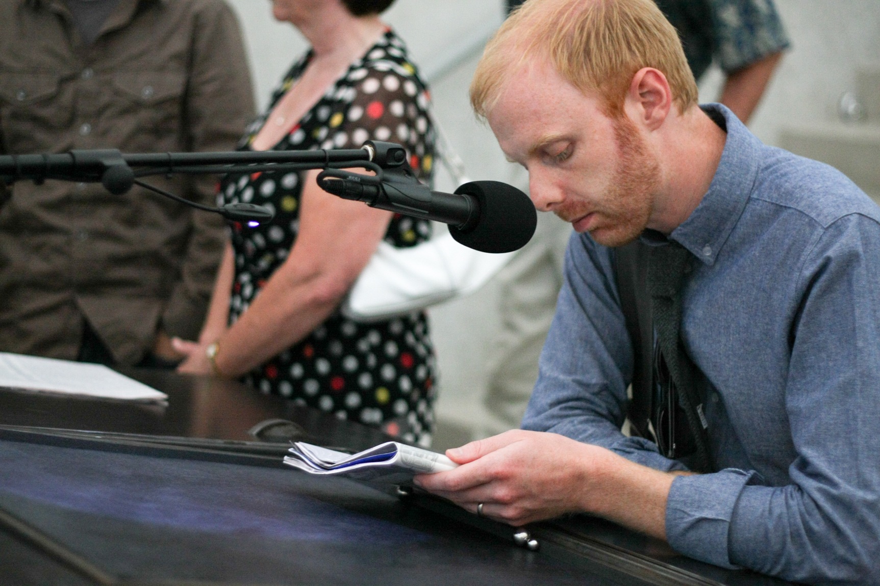 A visitor holds a book and leans against a table, a microphone in the foreground.