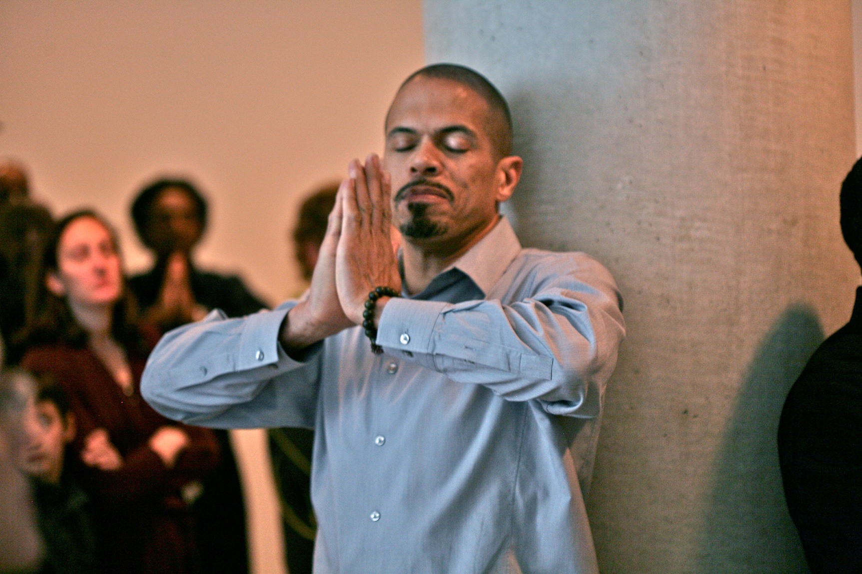 Participant raises hands in a praying pose with their back against a concrete pillar.