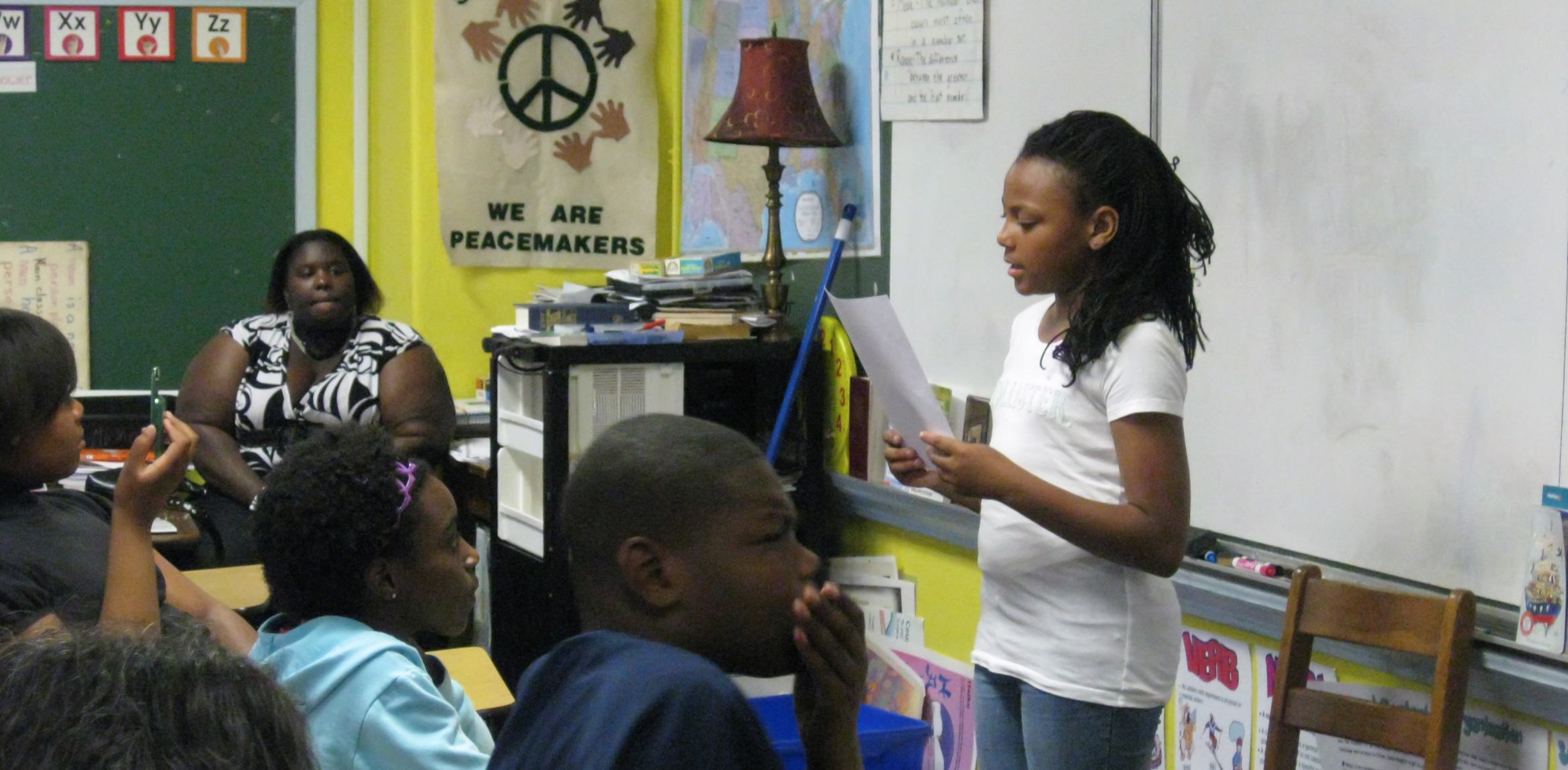 A student reads in front of a classroom of students and a teacher from a paper in their hands.