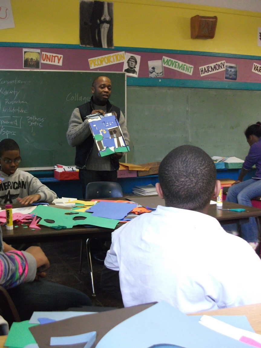 A teacher holds up a decorated piece of paper and speaks to a classroom of students.