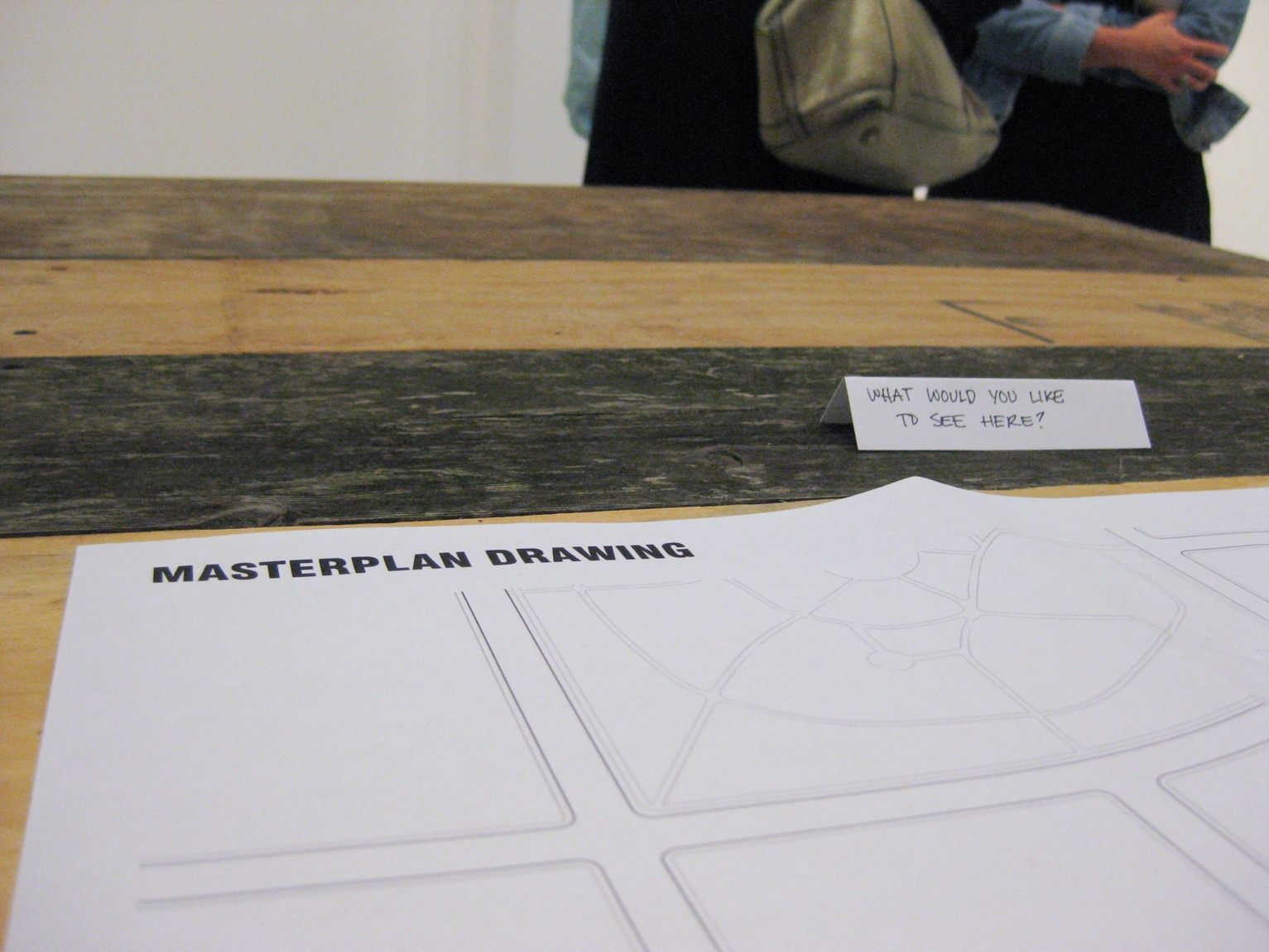 A closeup of a page that reads "MASTERPLAN DRAWING" and a small sign that reads "what would you like to see here?" lying on a wooden table.