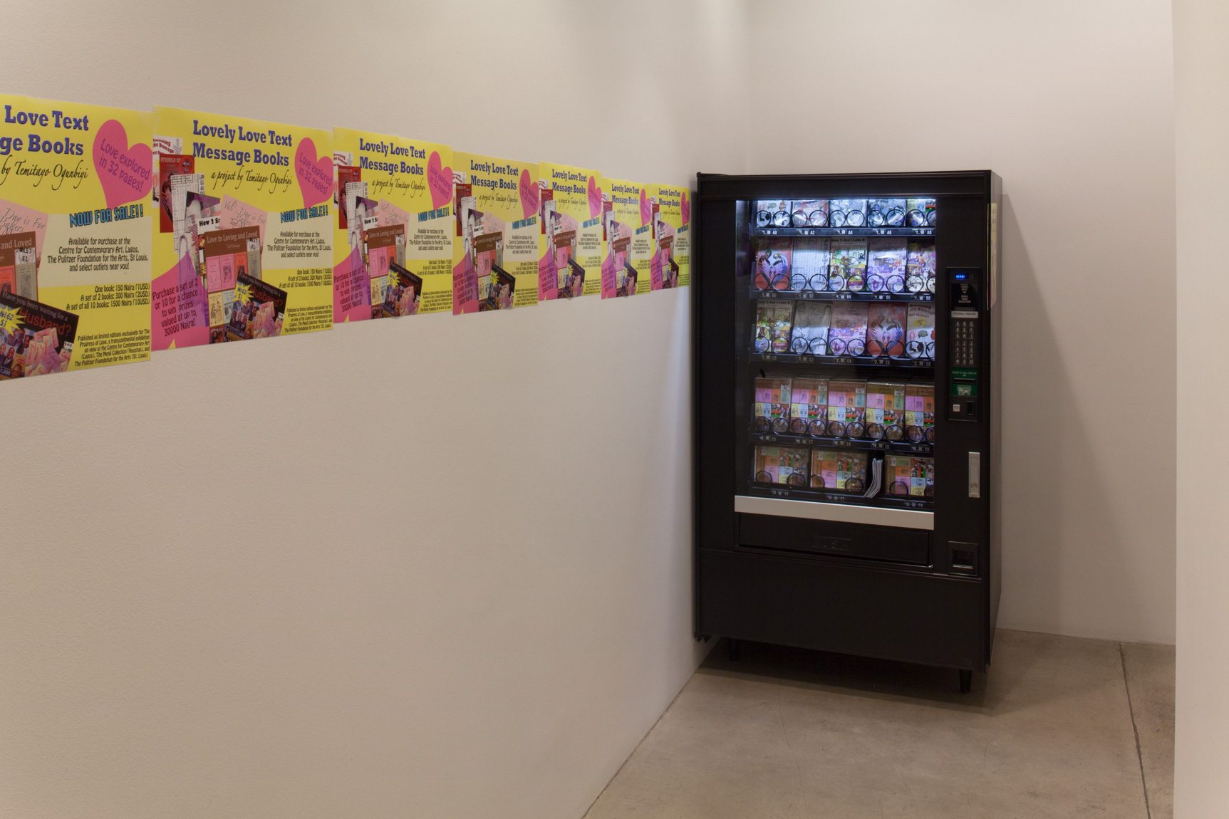 An exhibition view of Temitayo Ogunbiyi's piece "Lovely Love Text Message Books," which includes copies of a page arranged on the wall in a line leading to a vending machine.