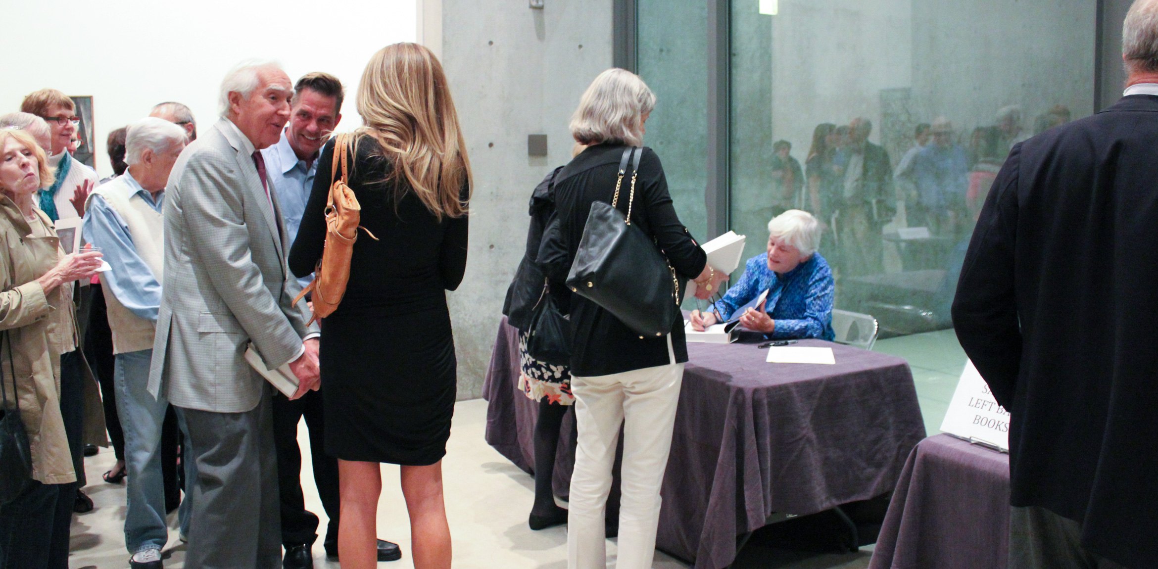 Marjorie B. Cohn chats with community members and signs their books at a table in front of the Water Court windows. Others gather to wait their turn to speak with her.