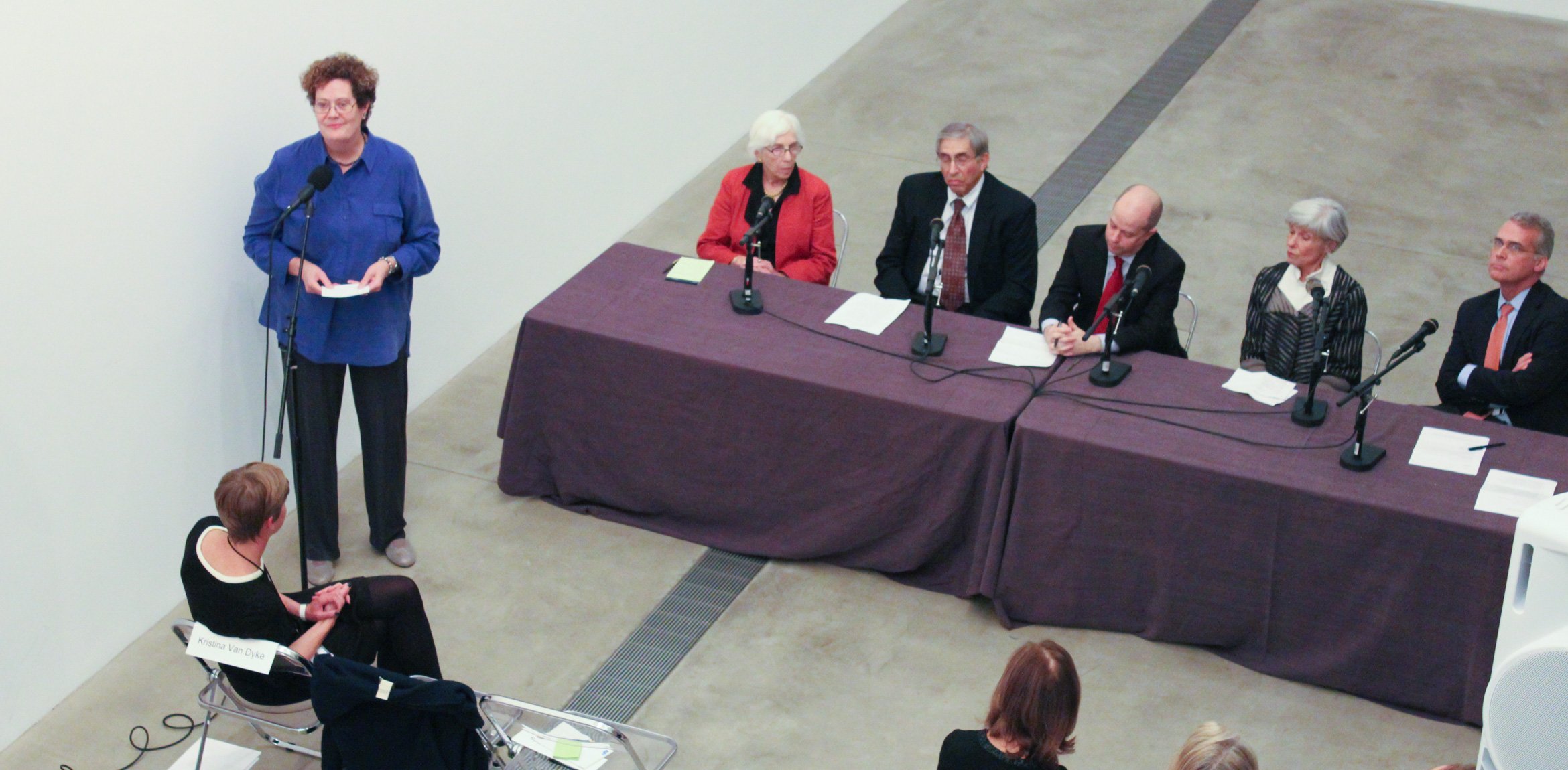 Elizabeth Childs stands at a microphone in the Lower-Main Gallery to the left of a panel table of collectors and looks at the audience.