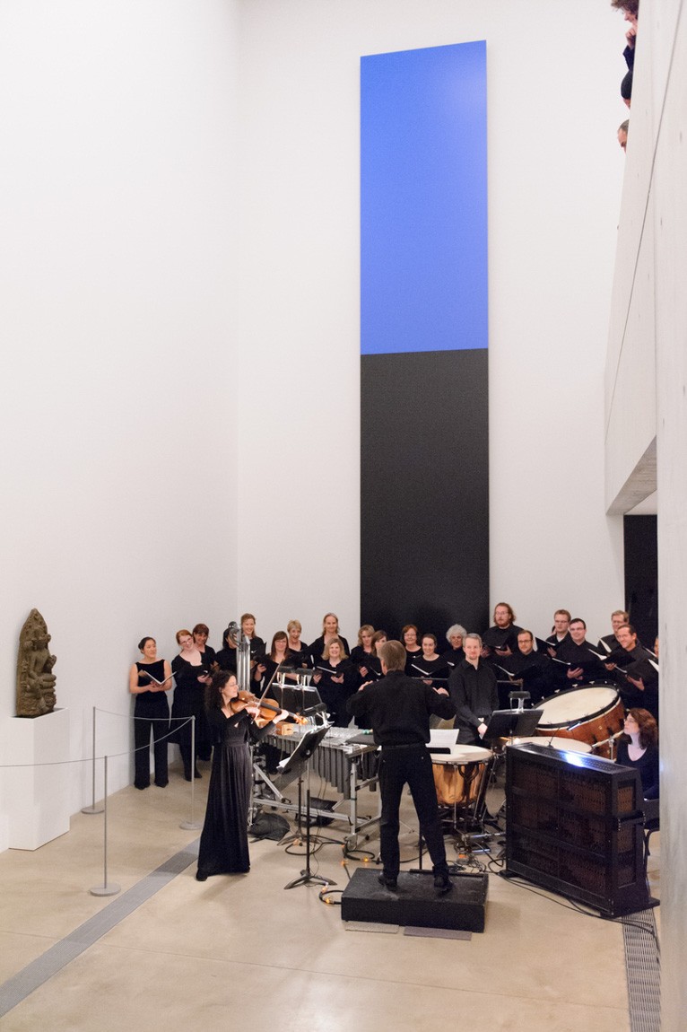 David Robertson conducts a violist and percussionist, and a choir group behind them in the Lower-Main Gallery.
