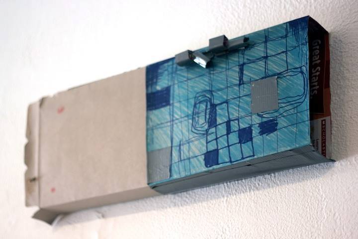 A closeup of a grey and blue cardboard box secured to the wall, with blue pen drawings on the blue side.