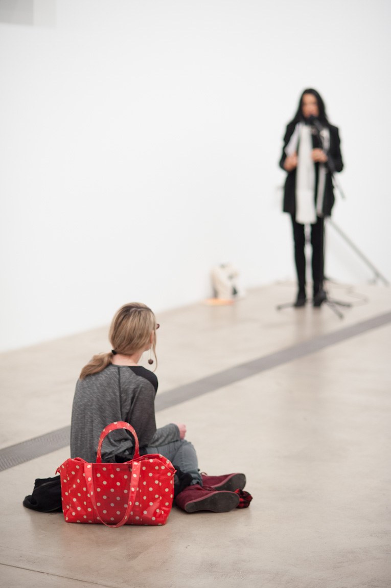 A lone visitor sits on the floor with a polka-dot red purse. In the background, Anne Waldman speaks into a microphone.