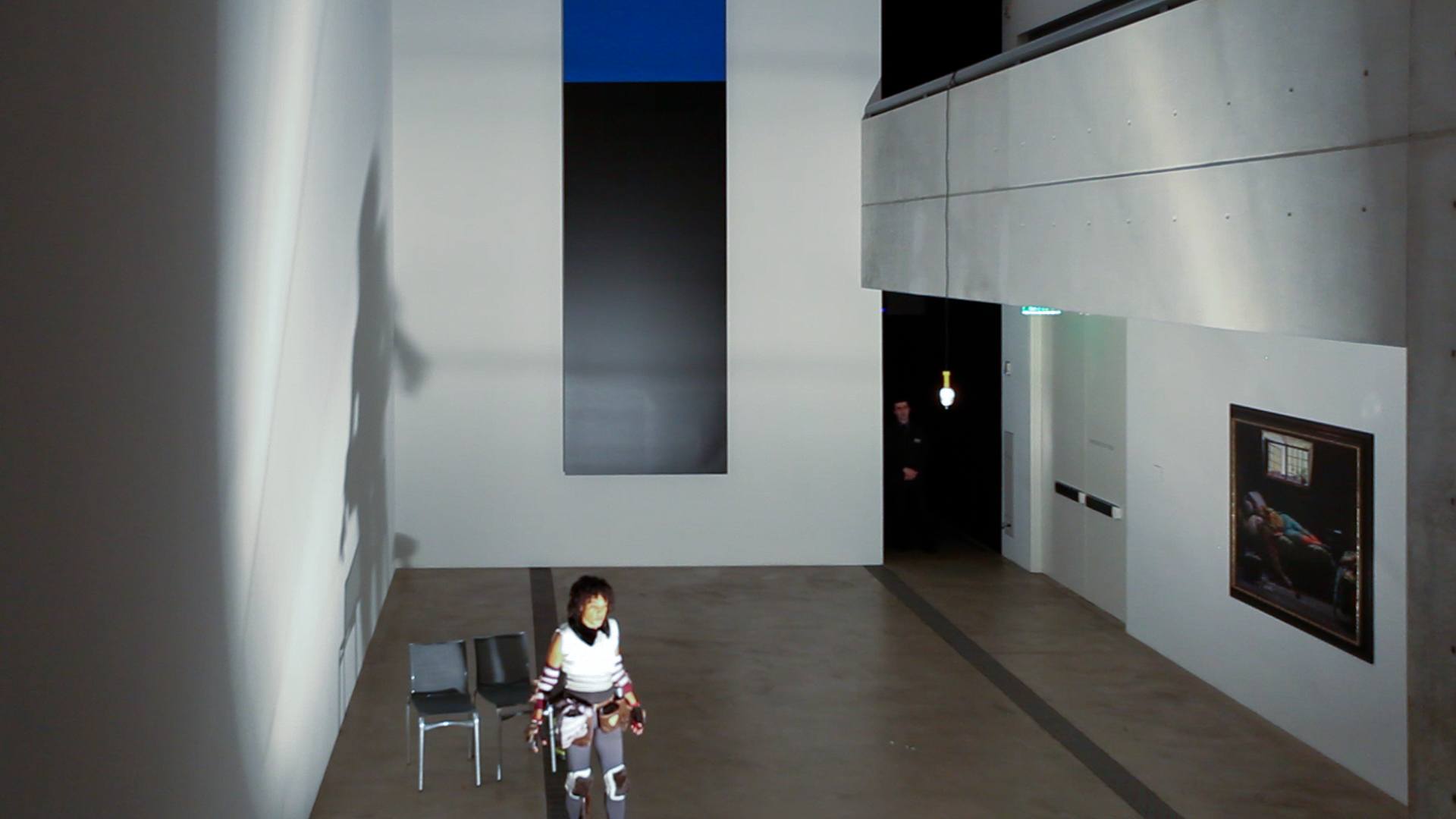 Wura-Natasha stands in the Lower-Main Gallery facing a bright light that casts her shadow on the wall.
