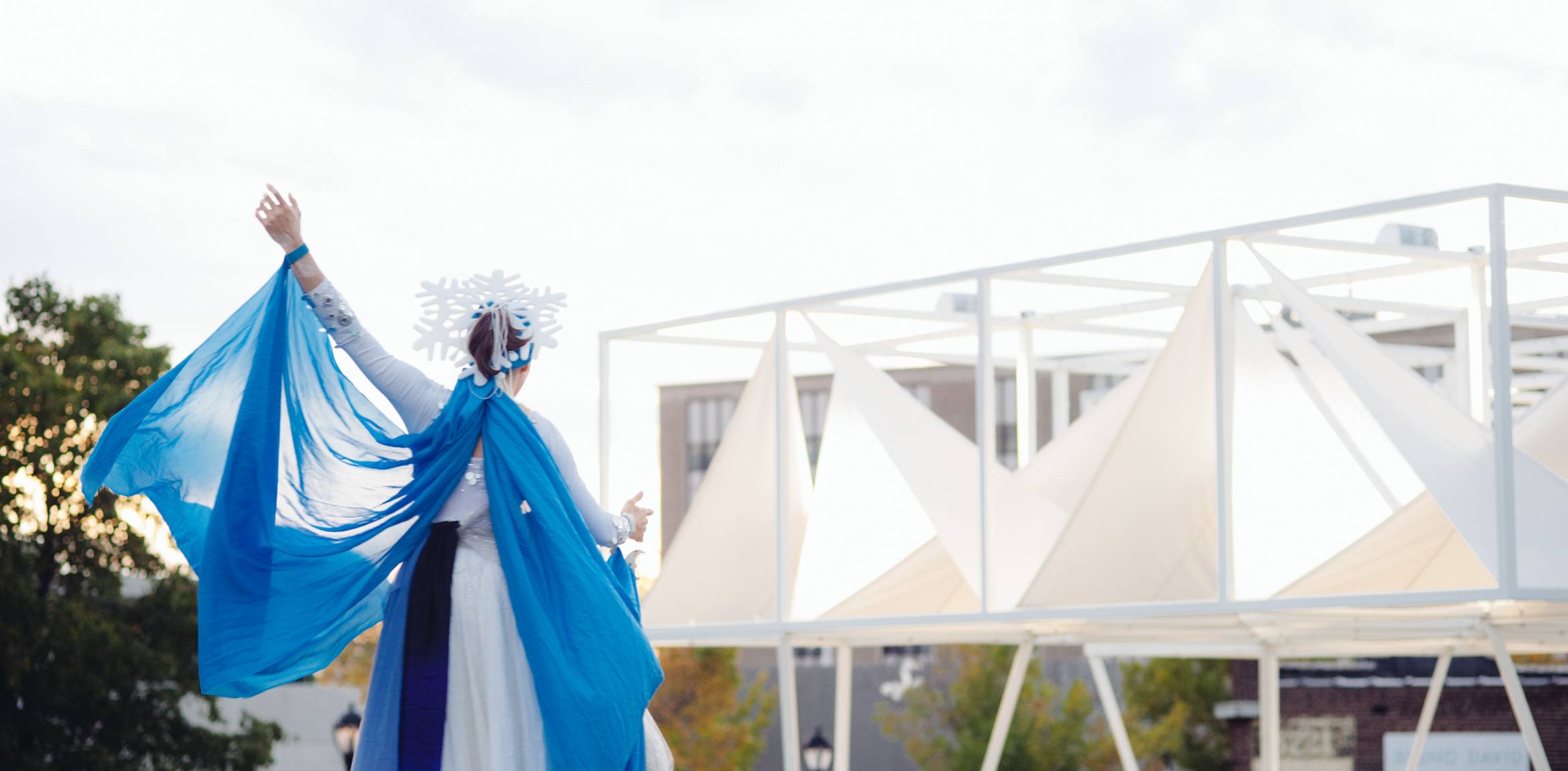 A performer wearing a white and blue flowing outfit with a snowflake headpiece raises an arm and dances, the Lots installation in the background.