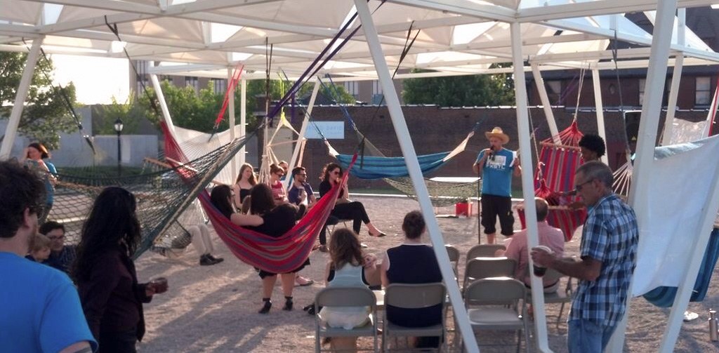 A group gathers under the Lots installation to listen to an individual wearing a cowboy hat who speaks into a microphone. Hammocks are strung and some visitors sit in them.