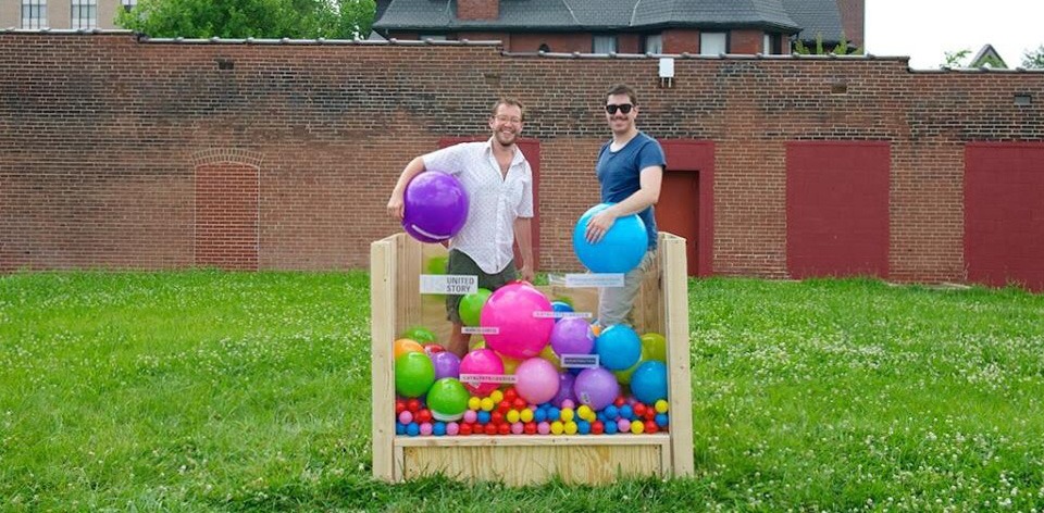 Two participants stand in a small glass ball pit in a grassy field in front of a brick wall. Each holds a large ball under an arm and smiles for the camera.