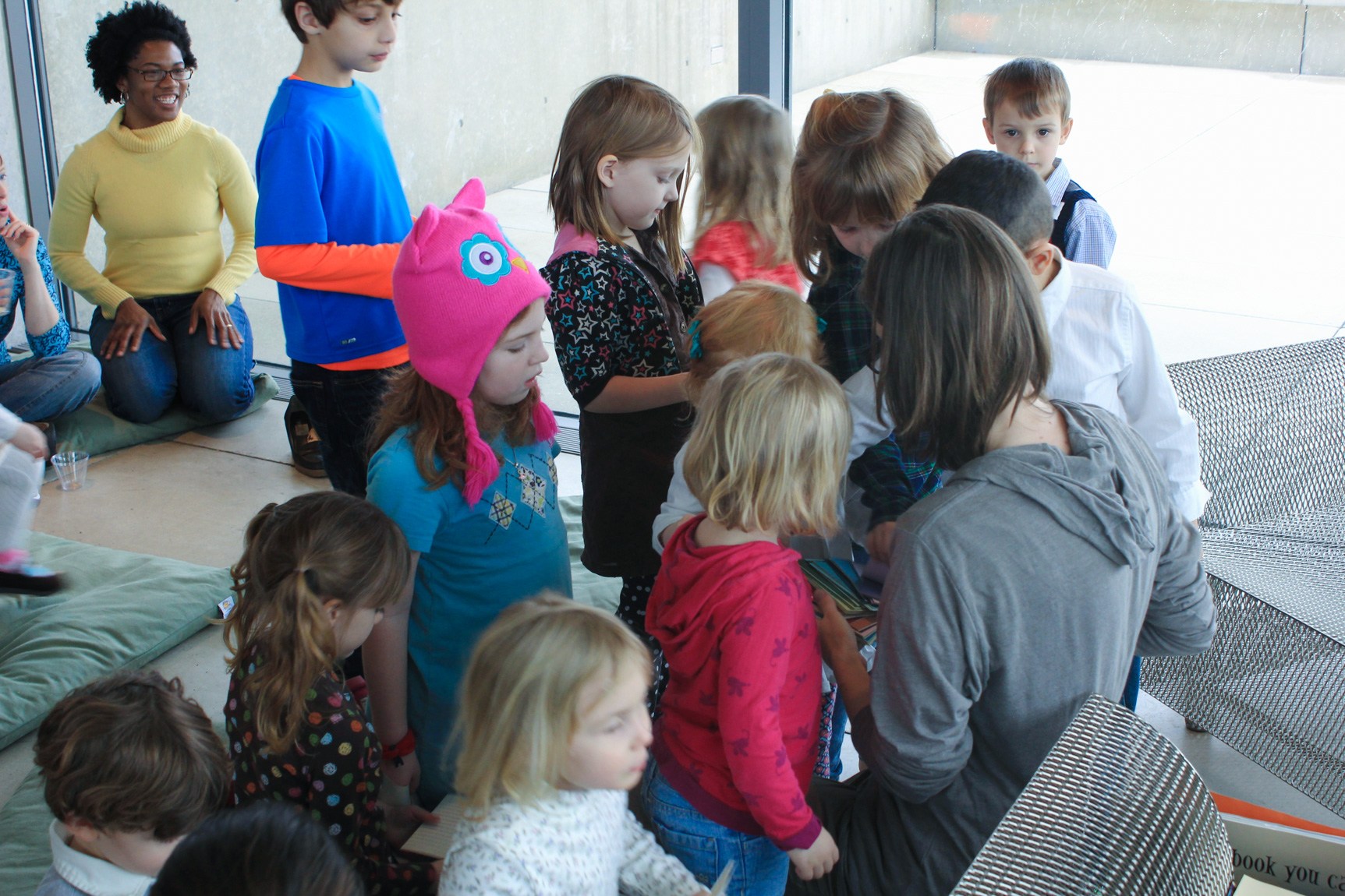 Children gather around a visitor reading a book in the Mezzanine while parents look on.