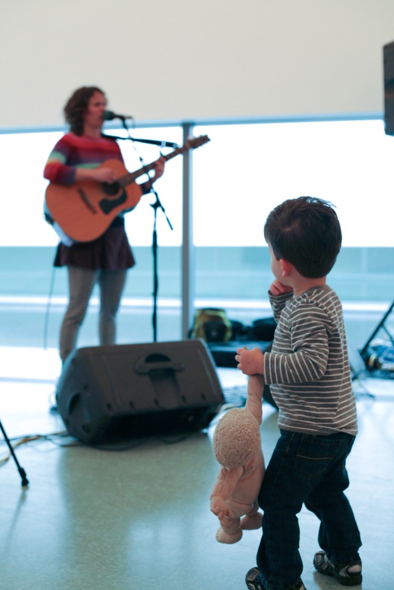 A child holds a stuffed animal and watches a musician sing and play guitar in the Main Gallery.