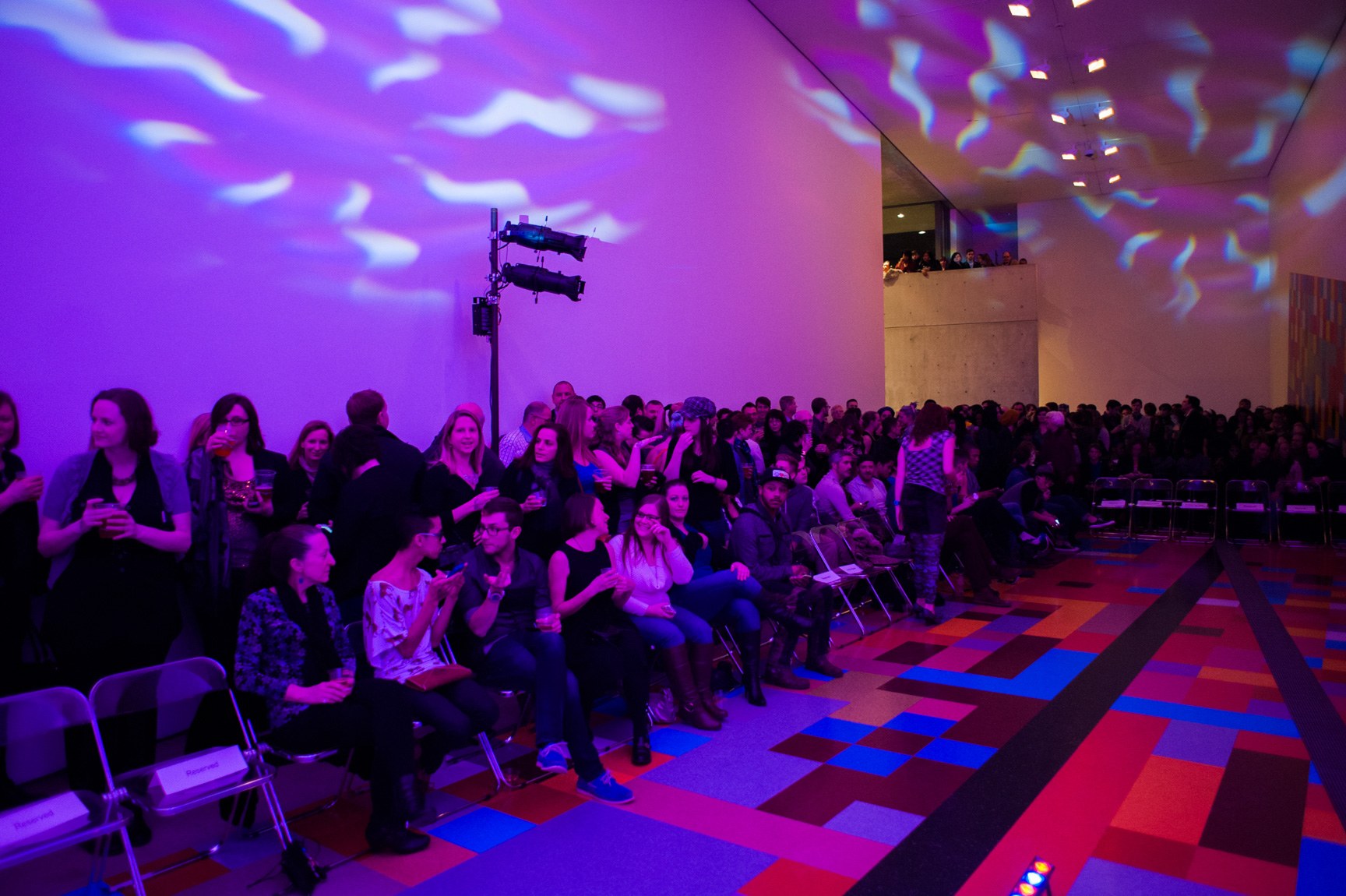 A large audience crowds the runway in the Main Gallery, bathed in purple and blue light.