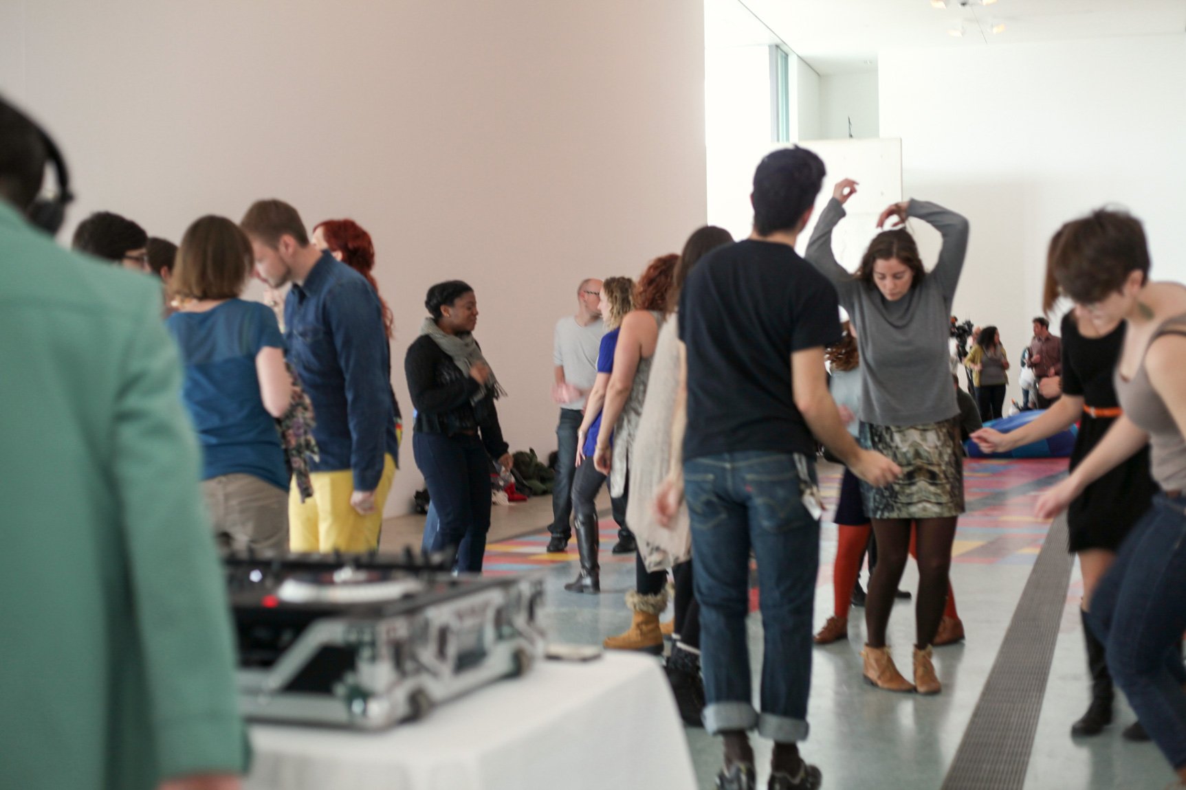 A large group of visitors dance in the Main Gallery while a DJ mixes at a turntable.