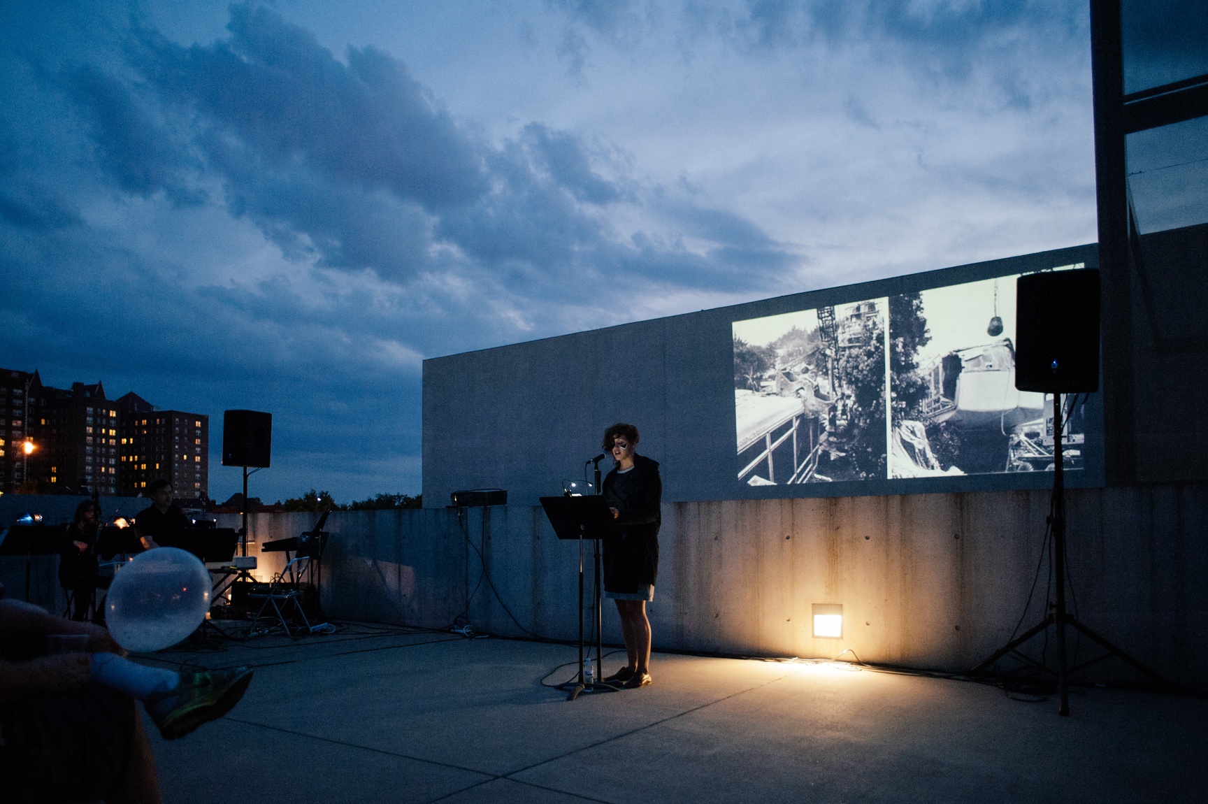During an evening event in the Courtyard, a presenter speaks into a microphone in front of a podium, with images projected on the concrete wall behind them.
