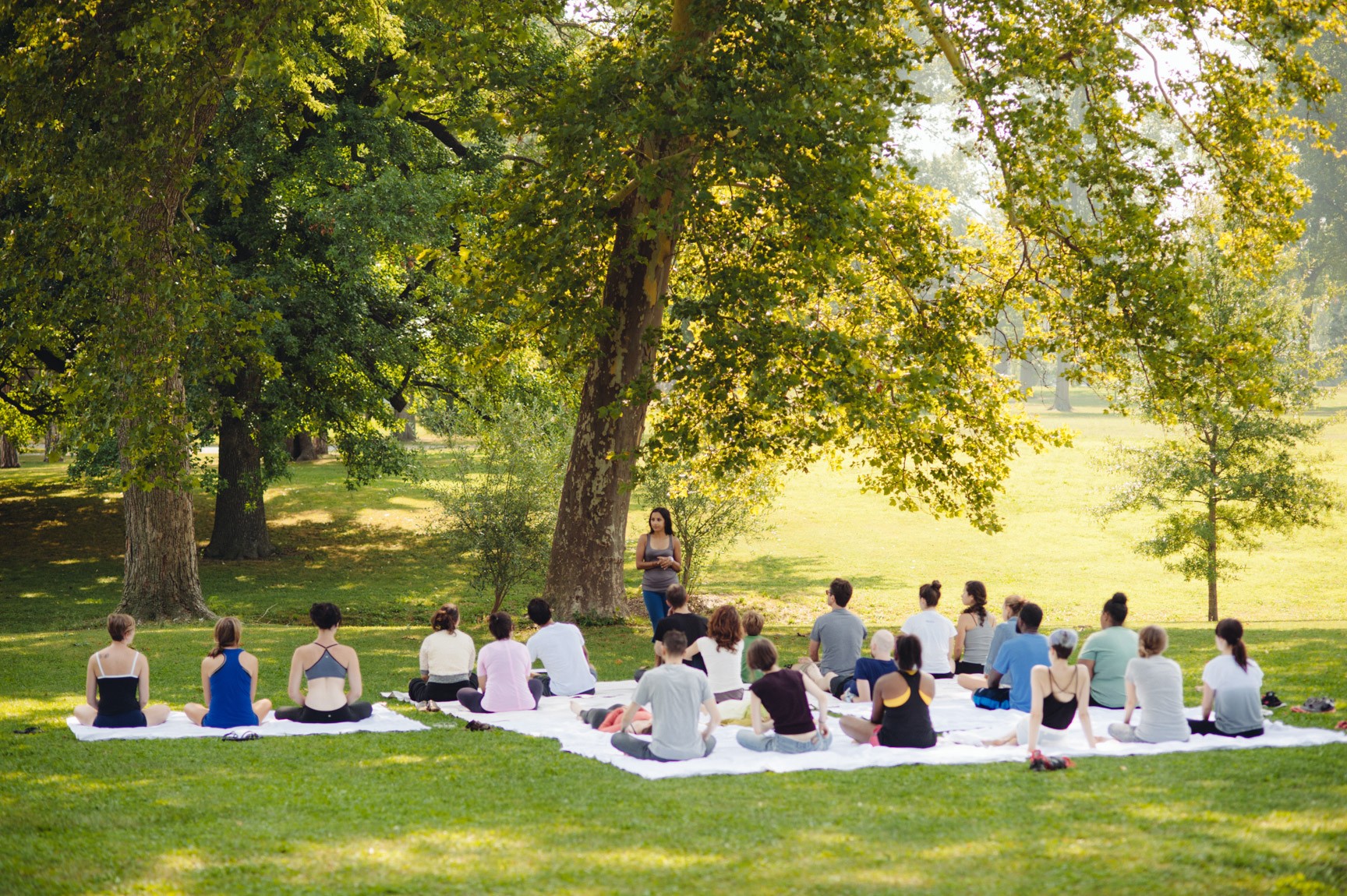 A large group is gathered on blankets in a shady field for guided meditation.