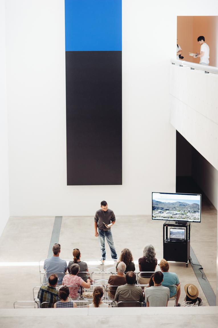 In the Lower-Main Gallery, a group sits in chairs in rows to watch a presenter speak with supplementary photos on a monitor.*what's the name?