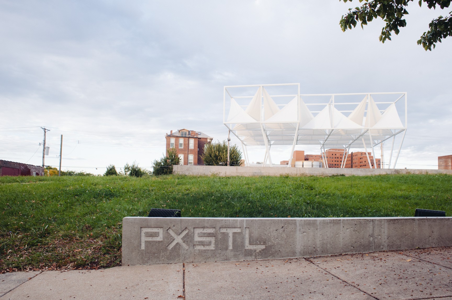 A clear view of the Lots installation, a geometric white sculpture with white triangular fabric pieces attached tautly to the structure's overhead beams. "PXSTL" is engraved into a concrete slab beside a sidewalk.
