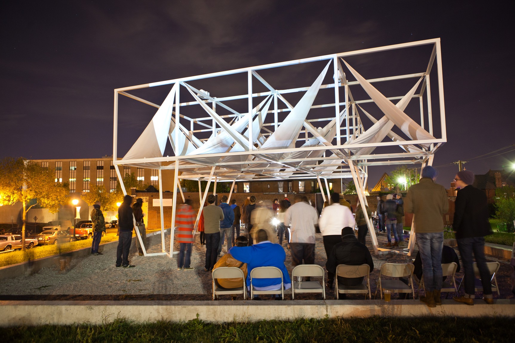 An audience gathers outdoors to watch performers under a large white installation canopy.