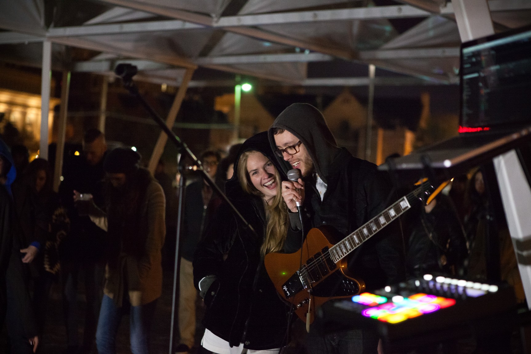 A friend smiles with Adult Fur, who speaks into a microphone and wears a hoodie and a guitar around his body.