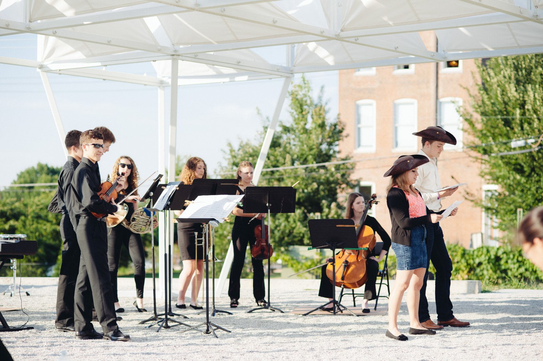 A group of student musicians in formal wear and two others wearing brown hats perform outdoors beneath a white geometric canopy installation.