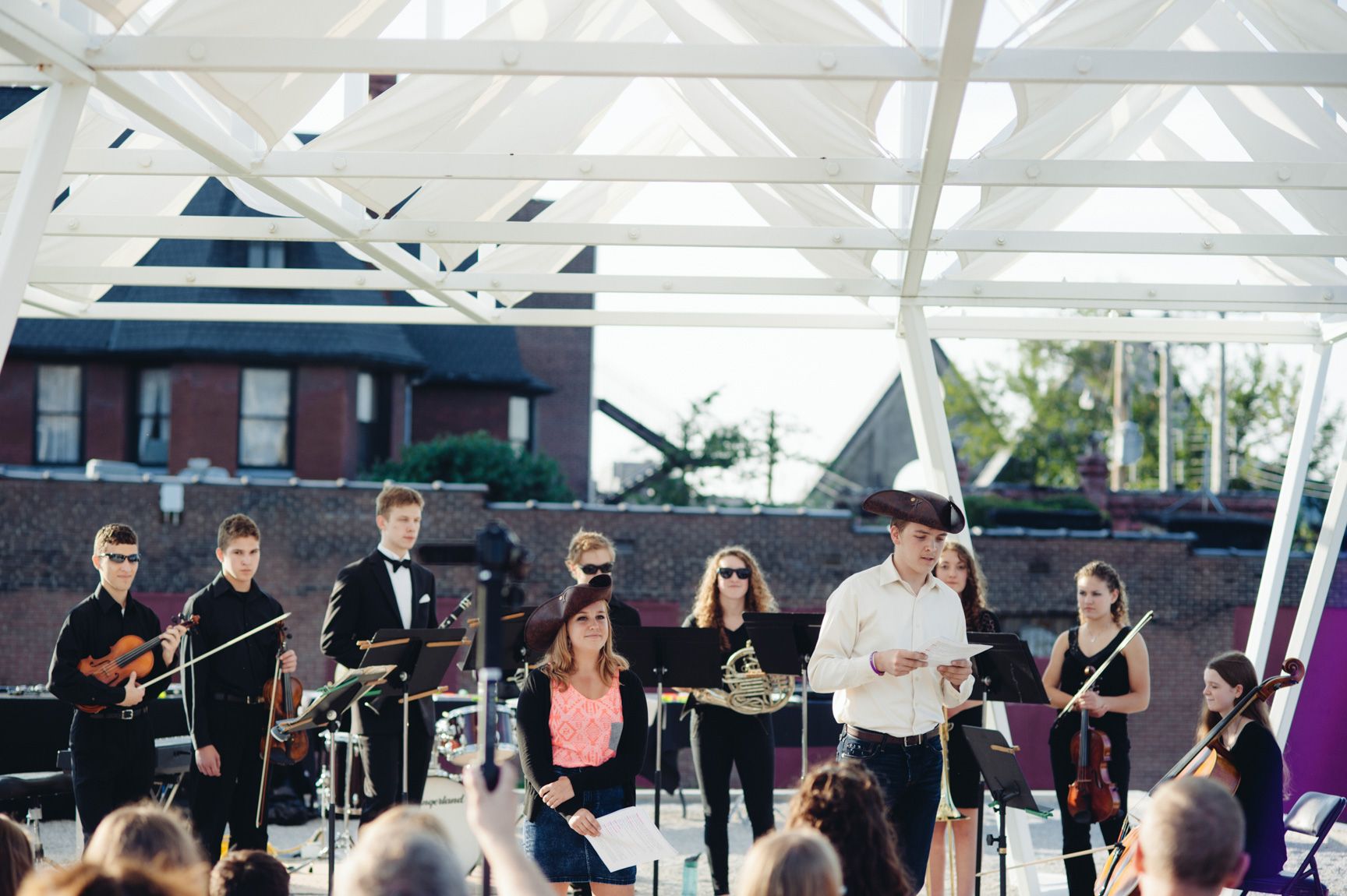 A group of student musicians in formal wear and two others wearing brown hats perform outdoors beneath a white geometric canopy installation for an audience.