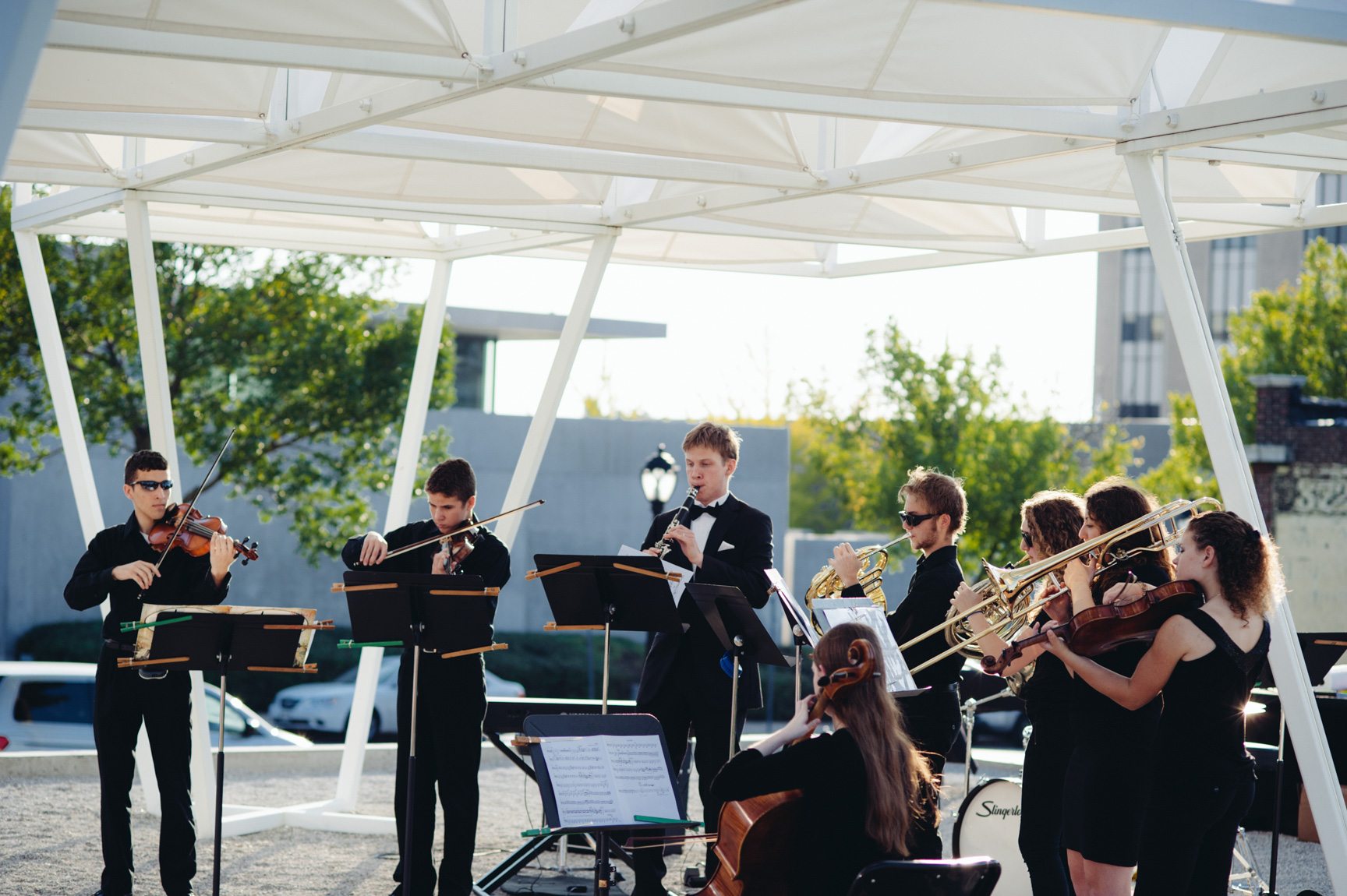 A group of student musicians in formal wear perform outdoors beneath a white geometric installation with a canopy.