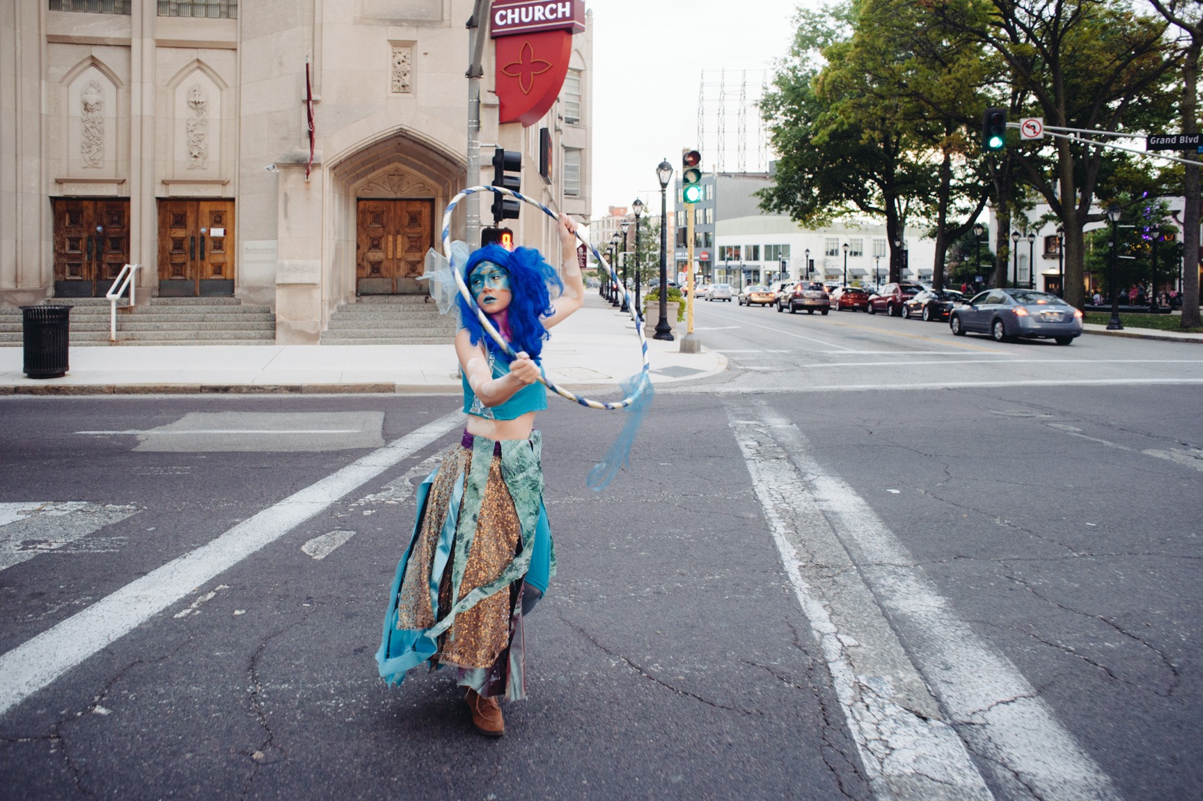 A performer wearing a blue wig and blue makeup carries a hula hoop above their head and crosses a street.