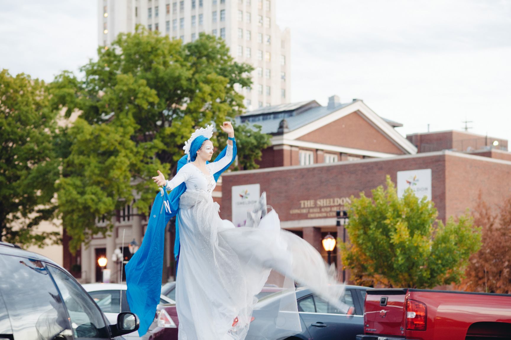 A performer dances on stilts outside, wearing a flowy white dress and a long blue scarf.