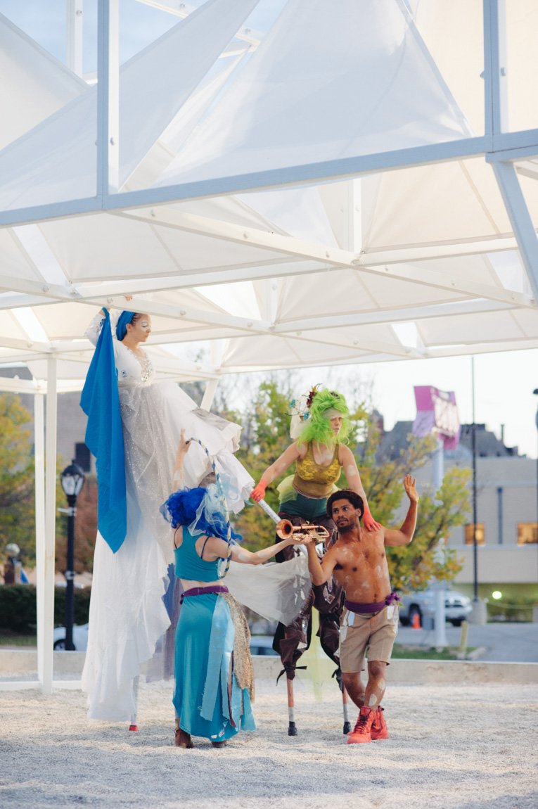 A small troupe wearing costumes performs beneath a large white installation canopy.