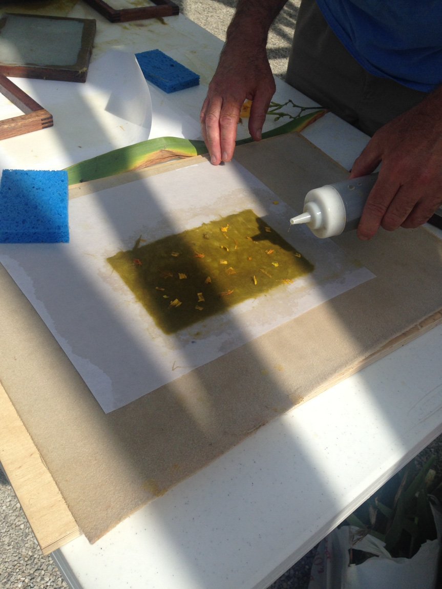 A visitor pours liquid out of a bottle onto a flat green piece of handmade paper on a worktable.