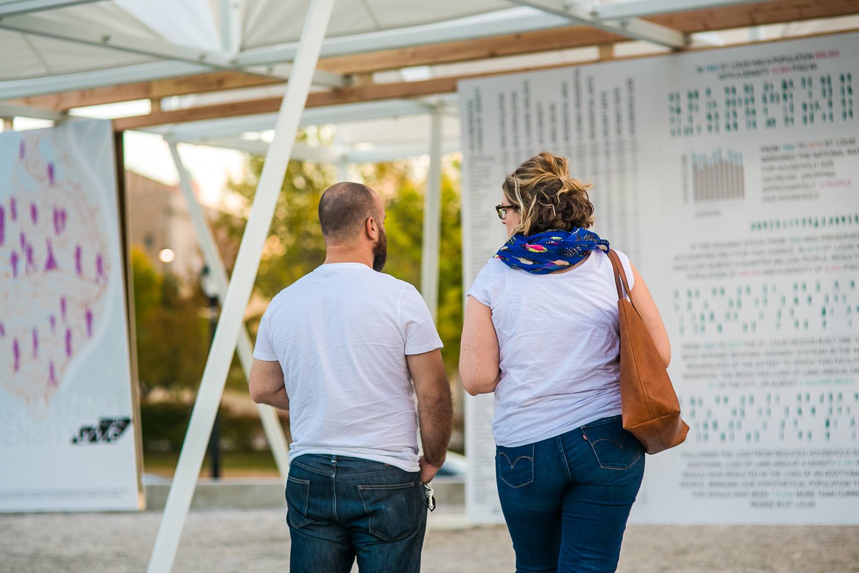 Two visitors stand before a wall of diagrams under a large white installation.