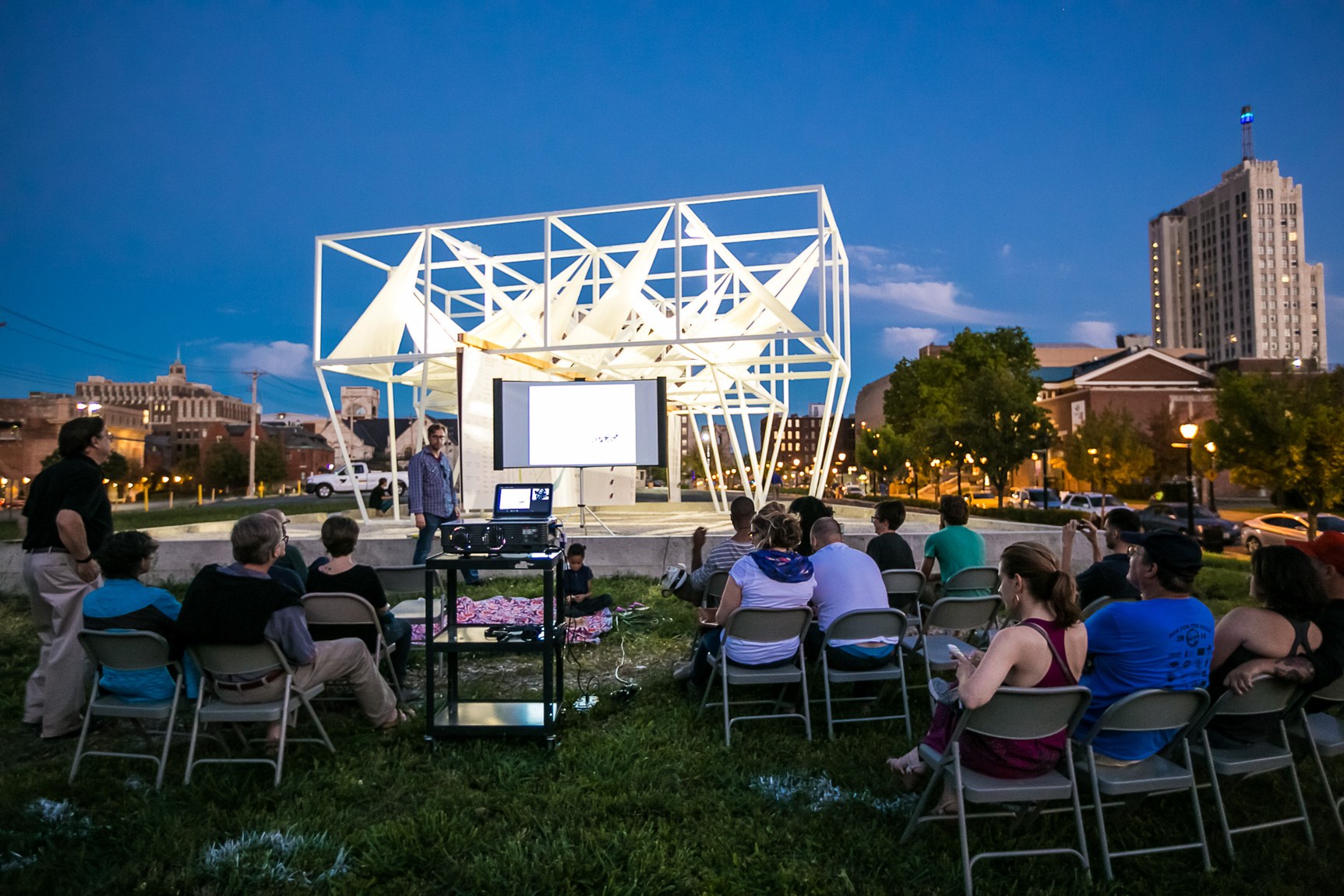 Brent Crittenden gives a lecture in front of a PowerPoint projected on a screen for an audience in front of a large white installation.
