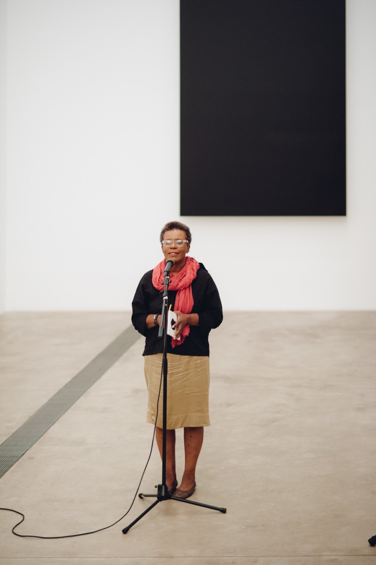 Claudia Rankine speaks into a microphone on a stand in front of "Blue Black" by Ellsworth Kelly.