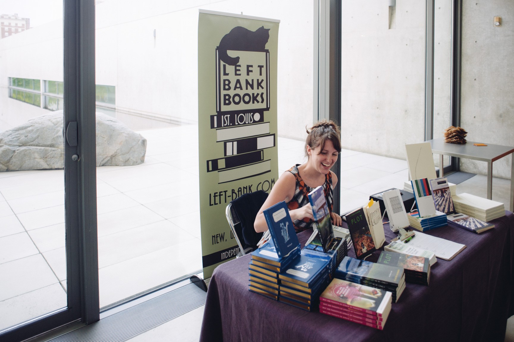 A vendor for Left Bank Books smiles down at their table of displayed literature in front of the Water Court windows.