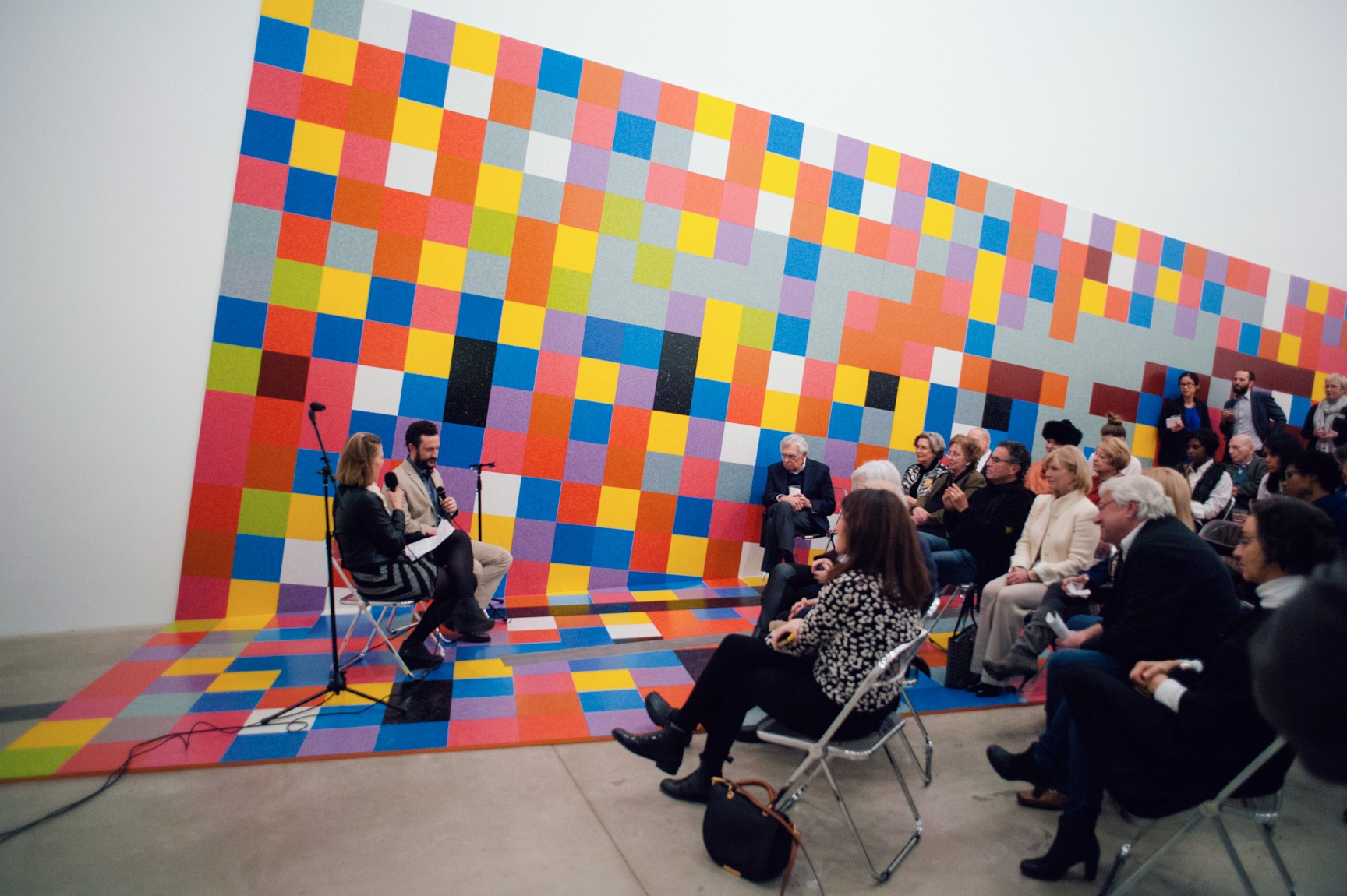 David Scanavino and an interviewer sit and speak into microphones for a seated audience in the Main Gallery, on Scanavino's colorful gridded installation, "Candy Crush."