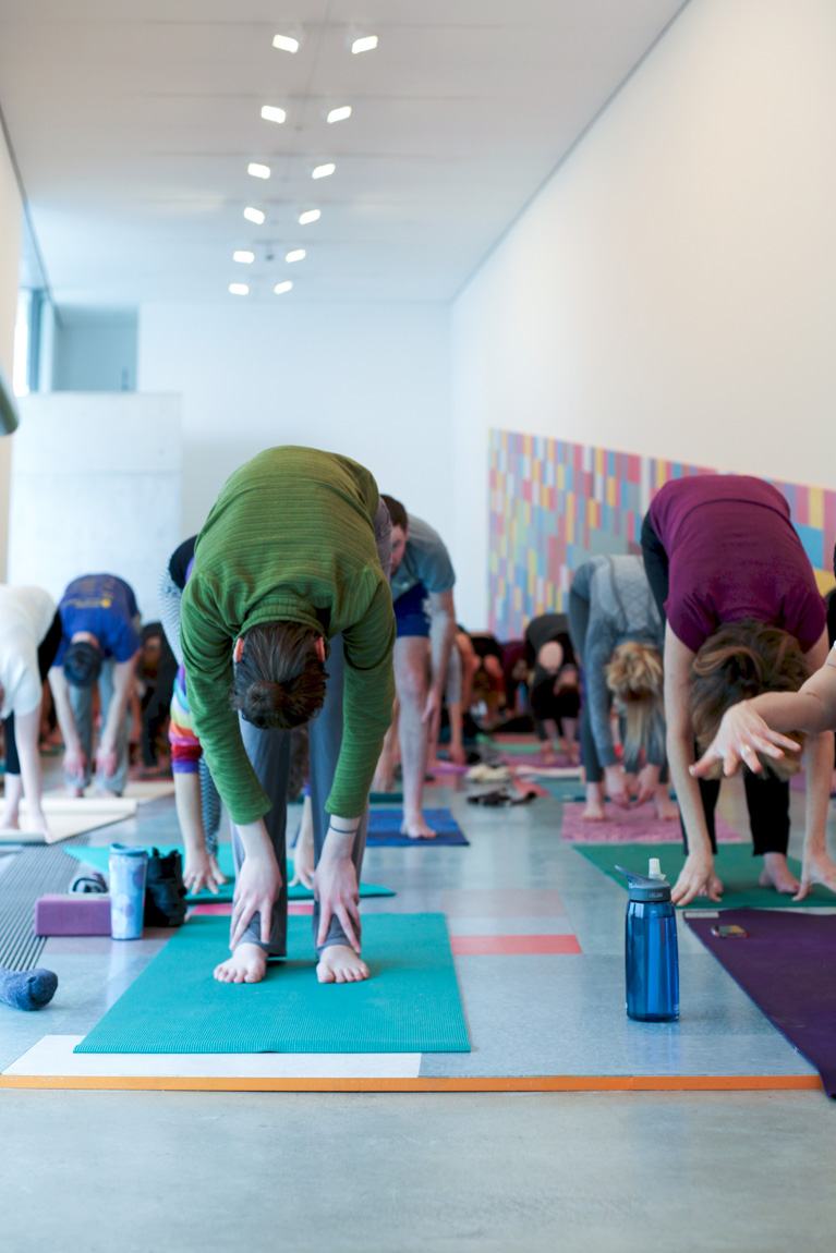 Yoga participants stand on yoga mats in the Main Gallery, bent over reaching their hands towards the floor.