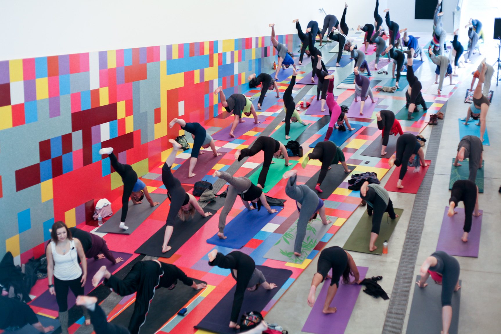 Laurie Brockhaus instructs yoga participants in the Main Gallery in downward dog poses, with their back leg lifted in the air.